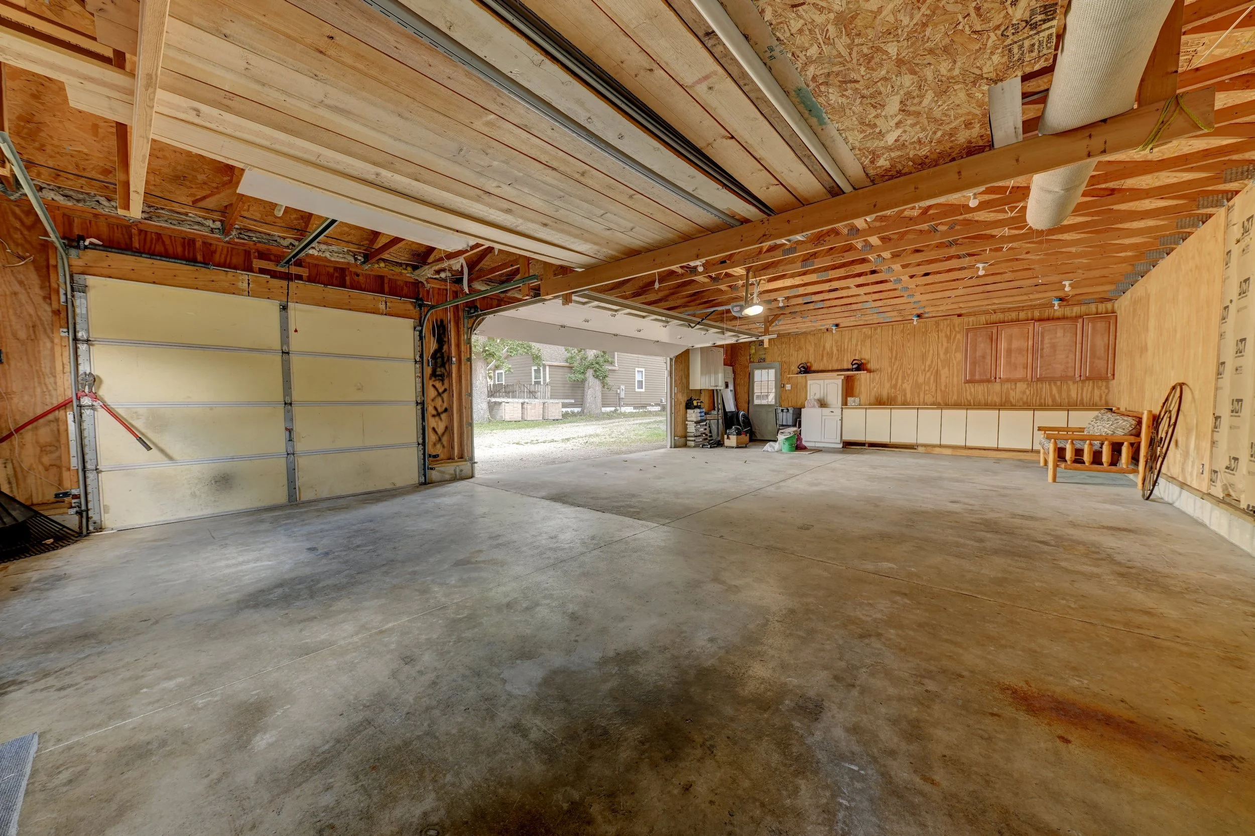 The interior of an unfinished garage with exposed wooden framing, a concrete floor, and an open garage door showing the outside yard with trees and neighboring houses.