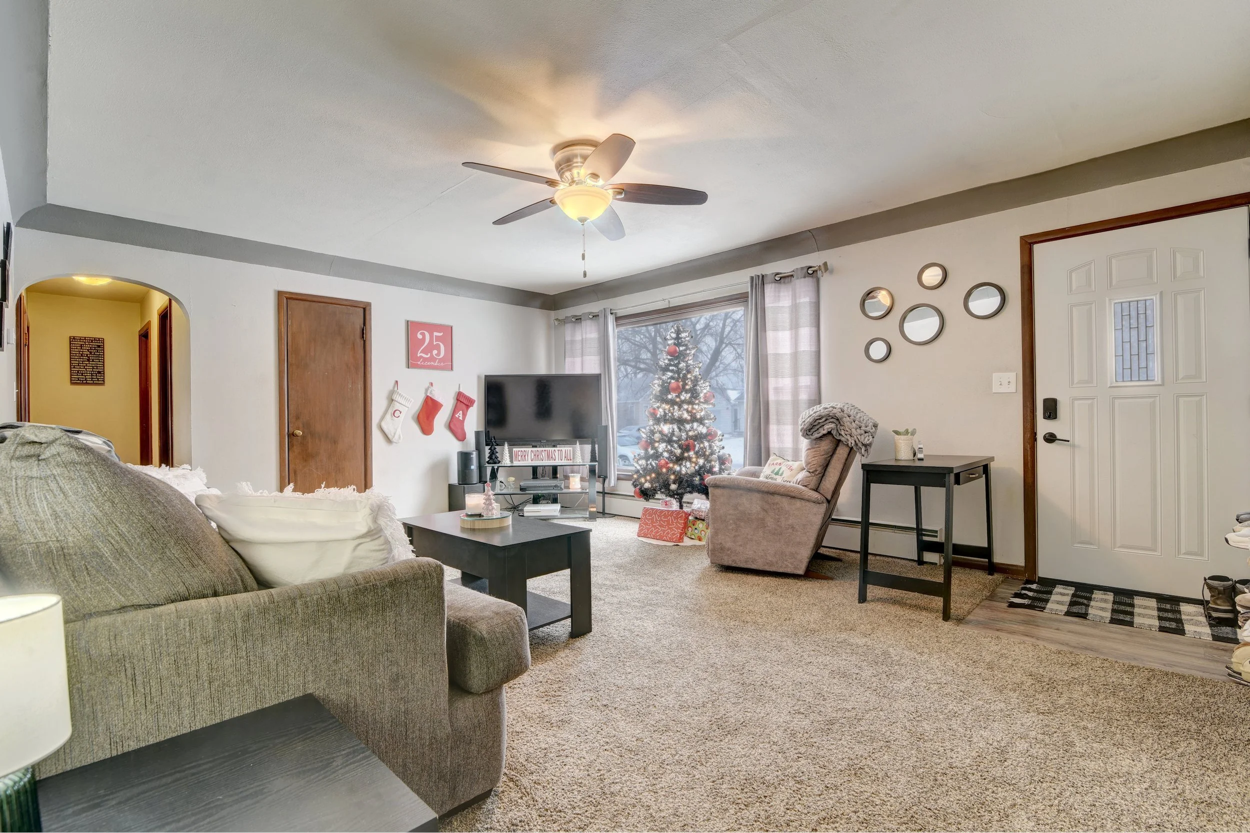 Living room decorated for Christmas with a Christmas tree, stockings on the wall, a beige recliner, a sofa with white pillows, a black coffee table, a TV, and a wall with circular mirrors.