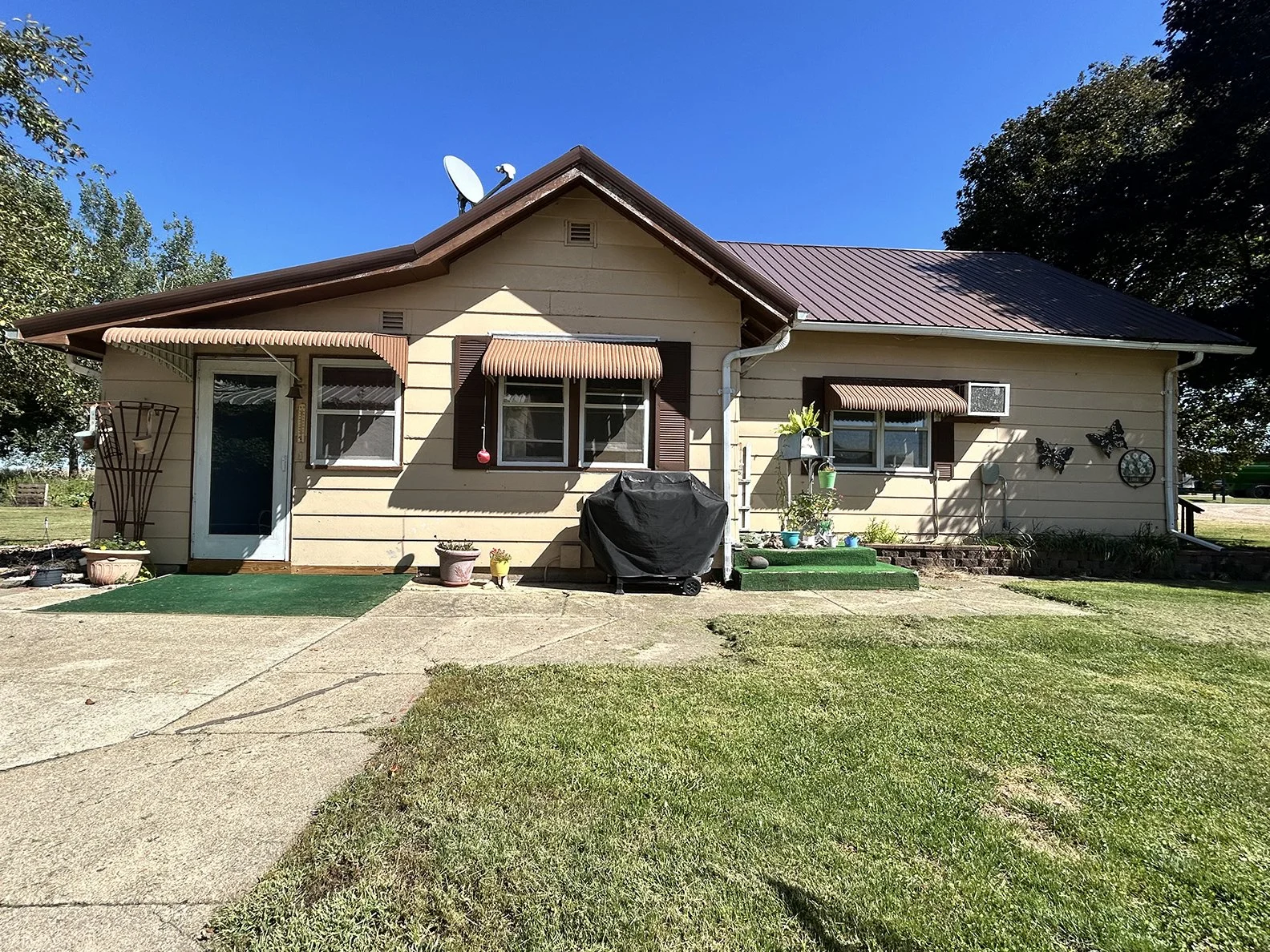 Front view of a beige house with brown shutters, awnings, and a metal roof. There is a green lawn, a concrete walkway, potted plants, a barbecue grill covered with a black cover, and decorative butterflies on the wall.