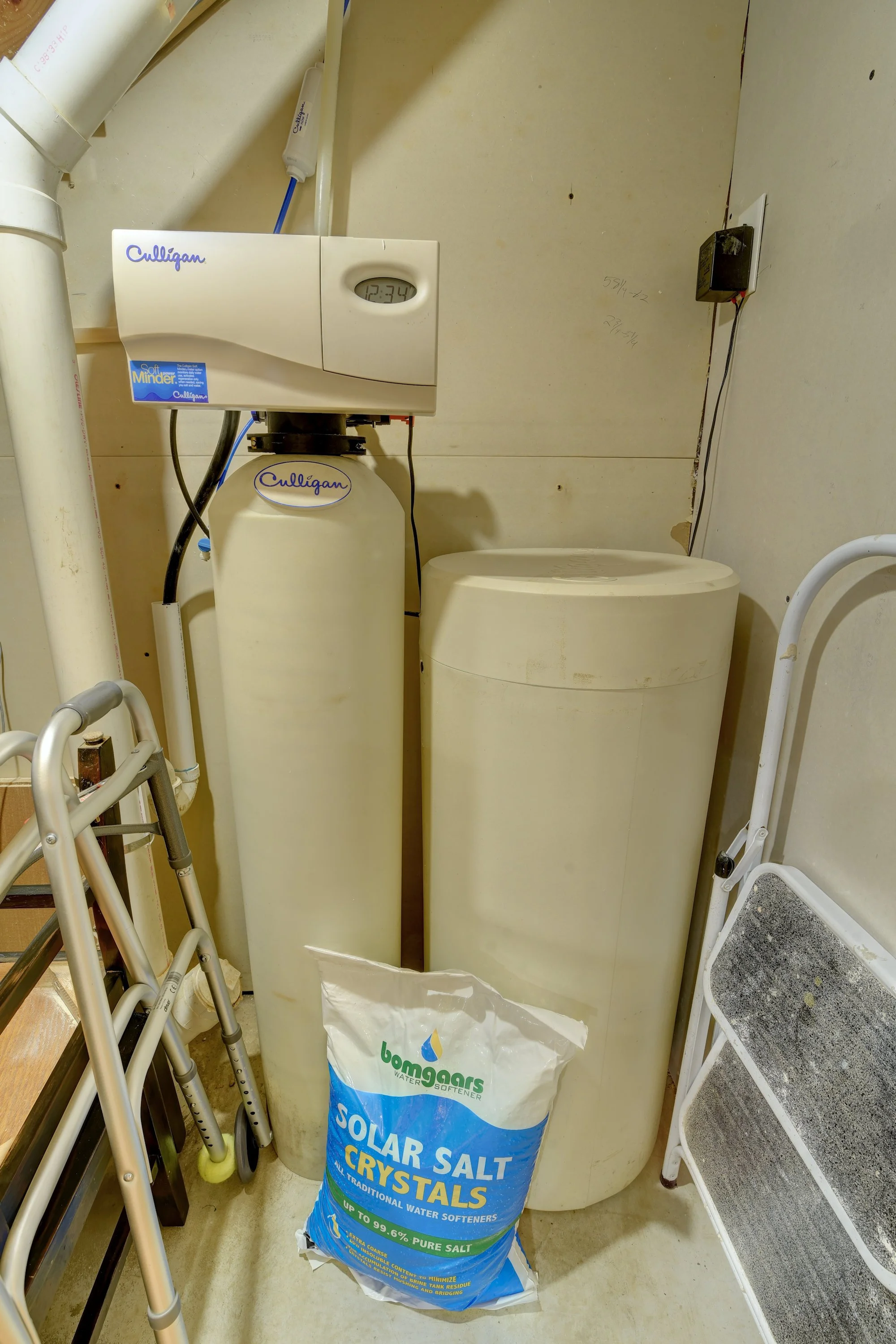 Water softener system with a Culligan control head, a large white resin tank, and a bag of solar salt crystals in front of the system, positioned in a utility room.