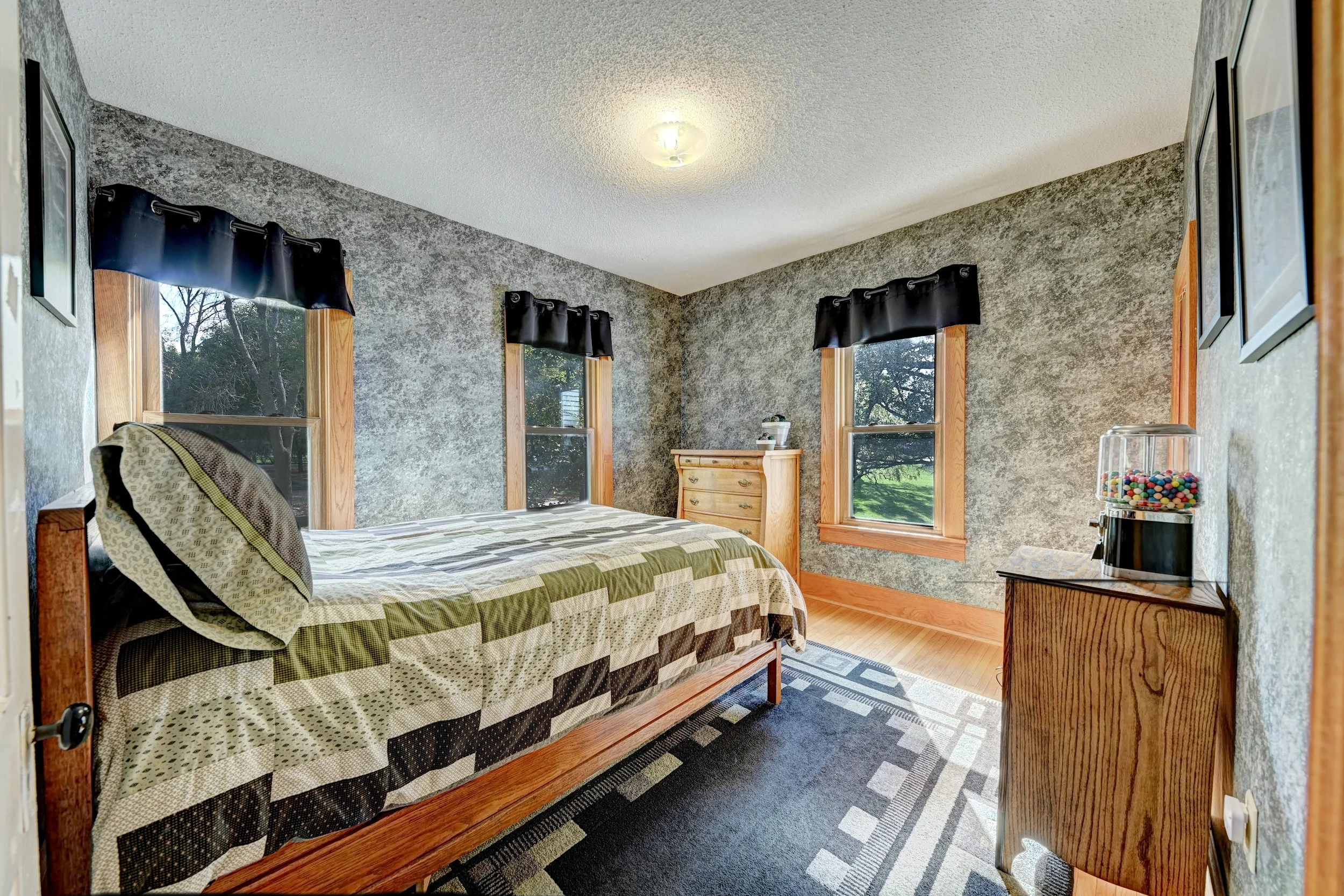 A bedroom with a wooden bed, green and brown plaid quilt, and a matching pillow. There are three windows with black valances and natural wood trim, a wooden dresser, a small wooden side table with a jar of colorful gumballs, and framed pictures on th