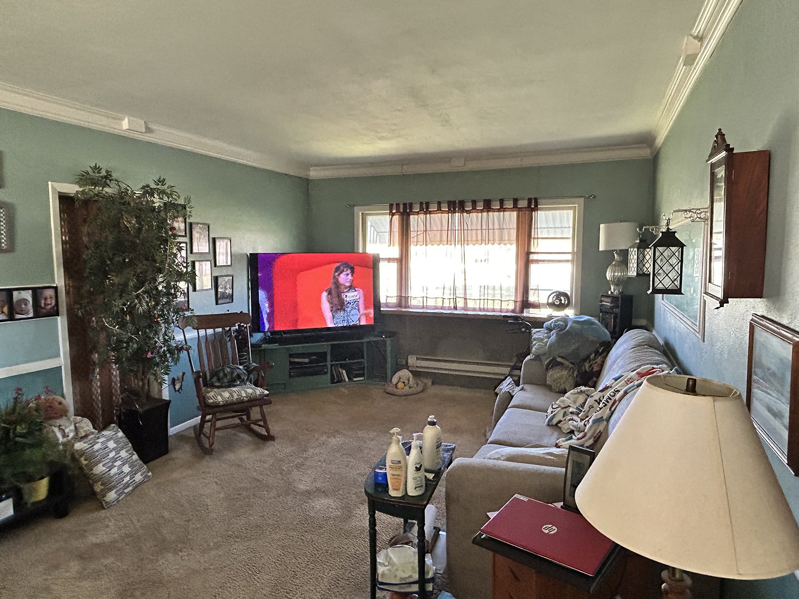 Living room with teal walls, a large window with curtains, a television on a stand showing a woman, a beige couch with blankets, various small furniture, pictures on the walls, a lamp, and a few bottles on a small table.