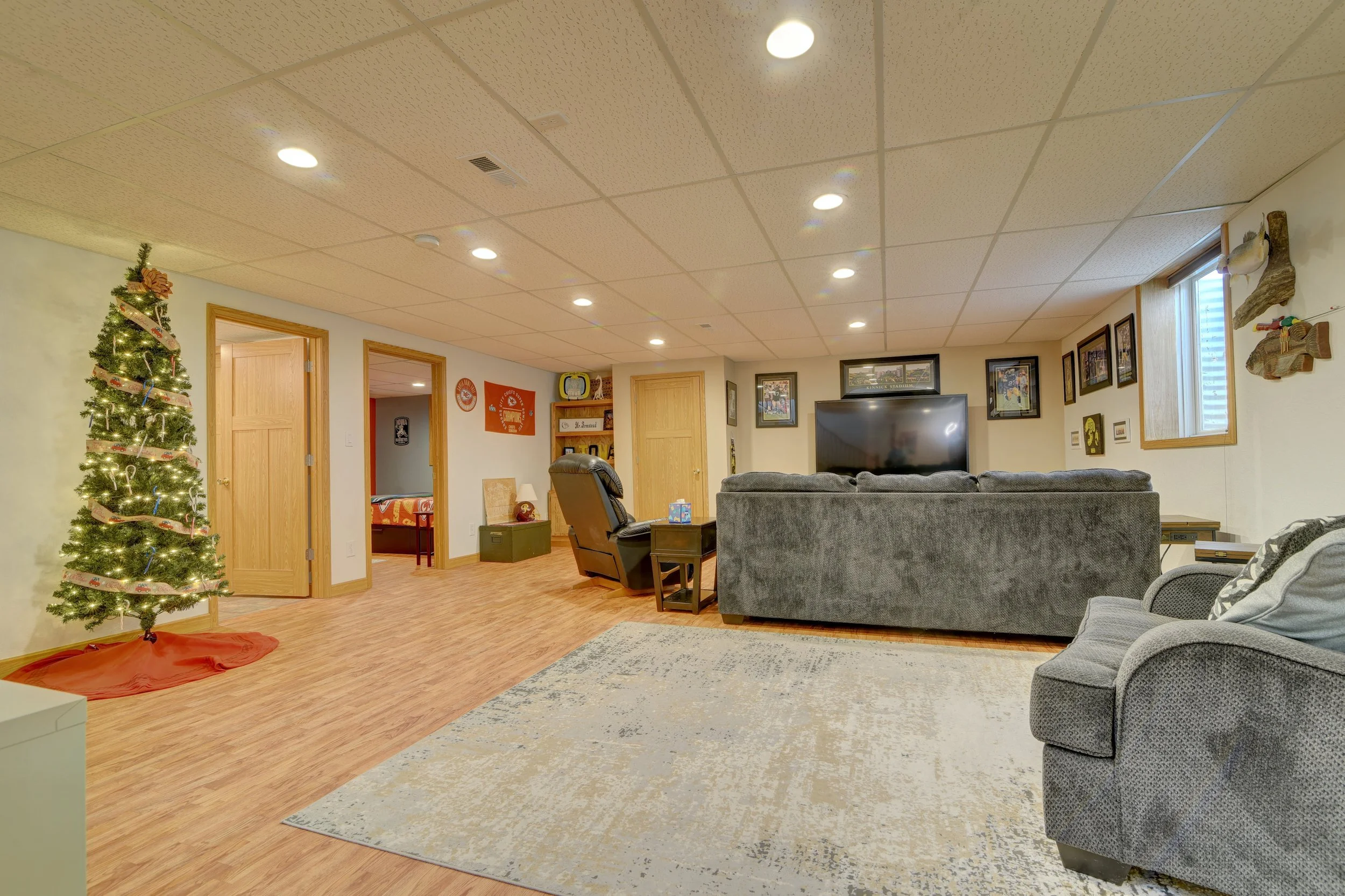Living room with a decorated Christmas tree on the left, a large wall-mounted TV on the far wall, gray sofas and armchair, framed pictures, and windows with blinds.