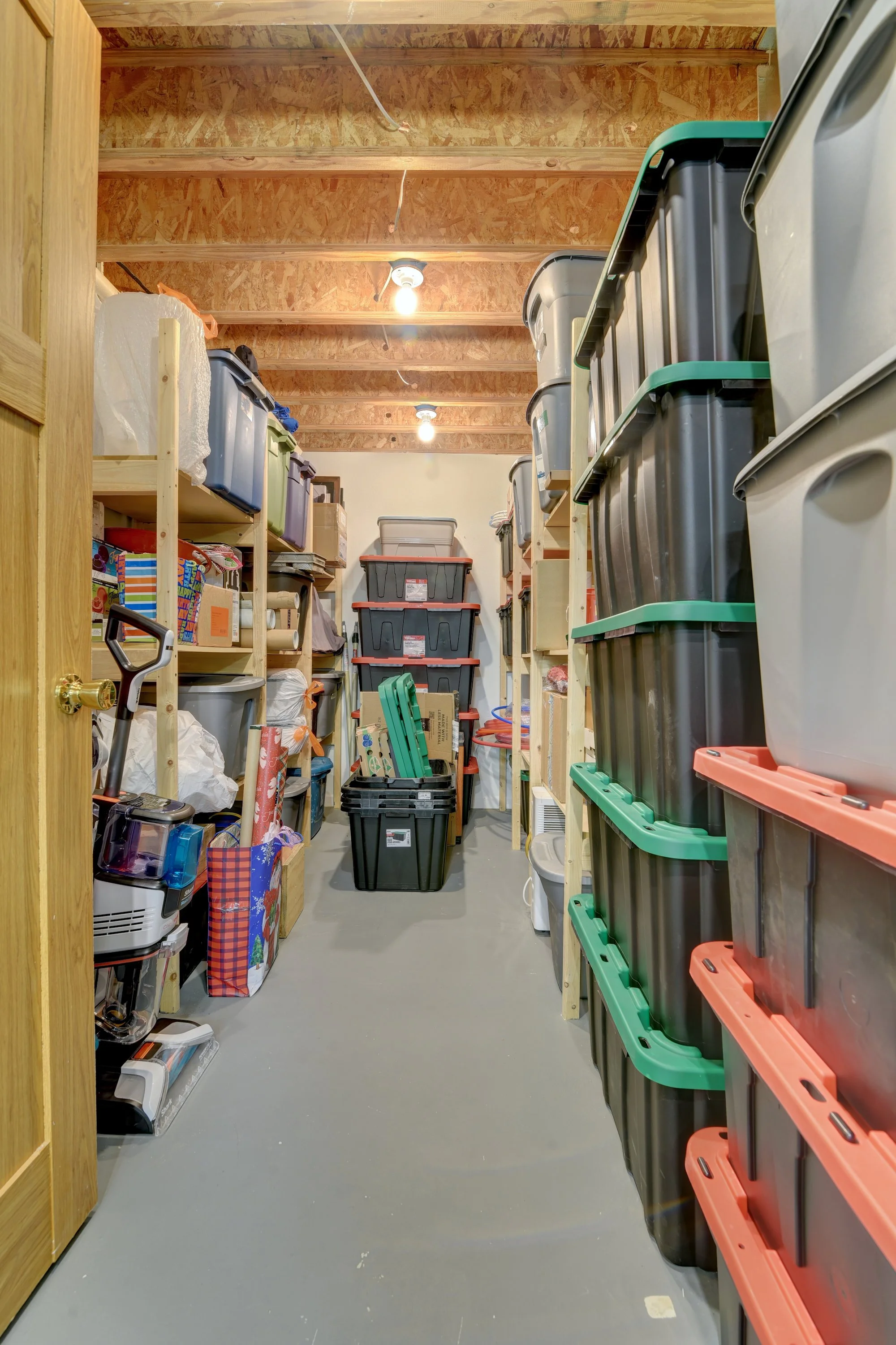 Storage room with wooden shelves filled with plastic storage bins and boxes, a shovel, and various household items, with a concrete floor and exposed wooden ceiling.