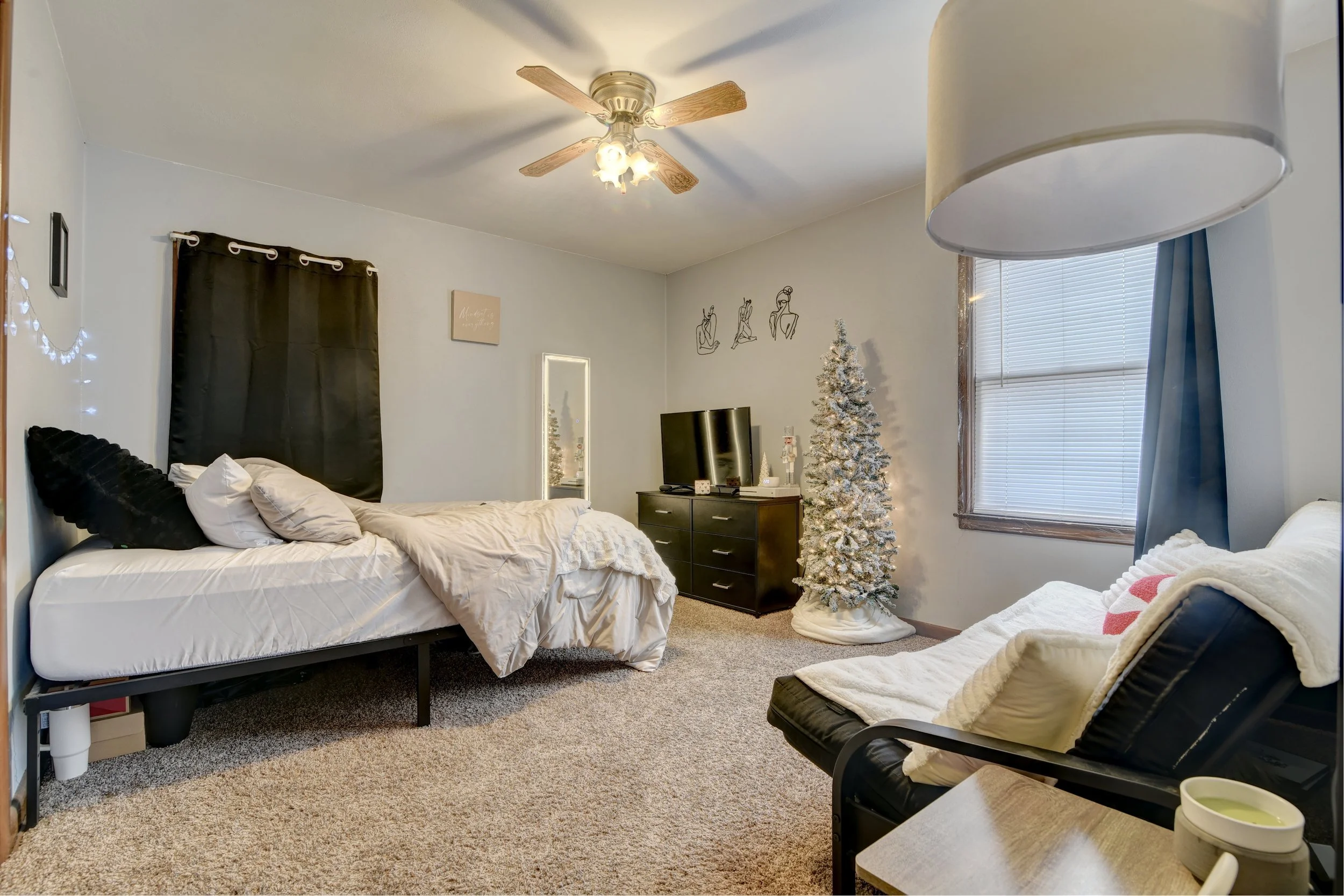 Bedroom with a bed, black curtains, a Christmas tree, a small black dresser with a TV, a mirror, a chair with a blanket, and window with blinds and curtain, decorated for the holidays.