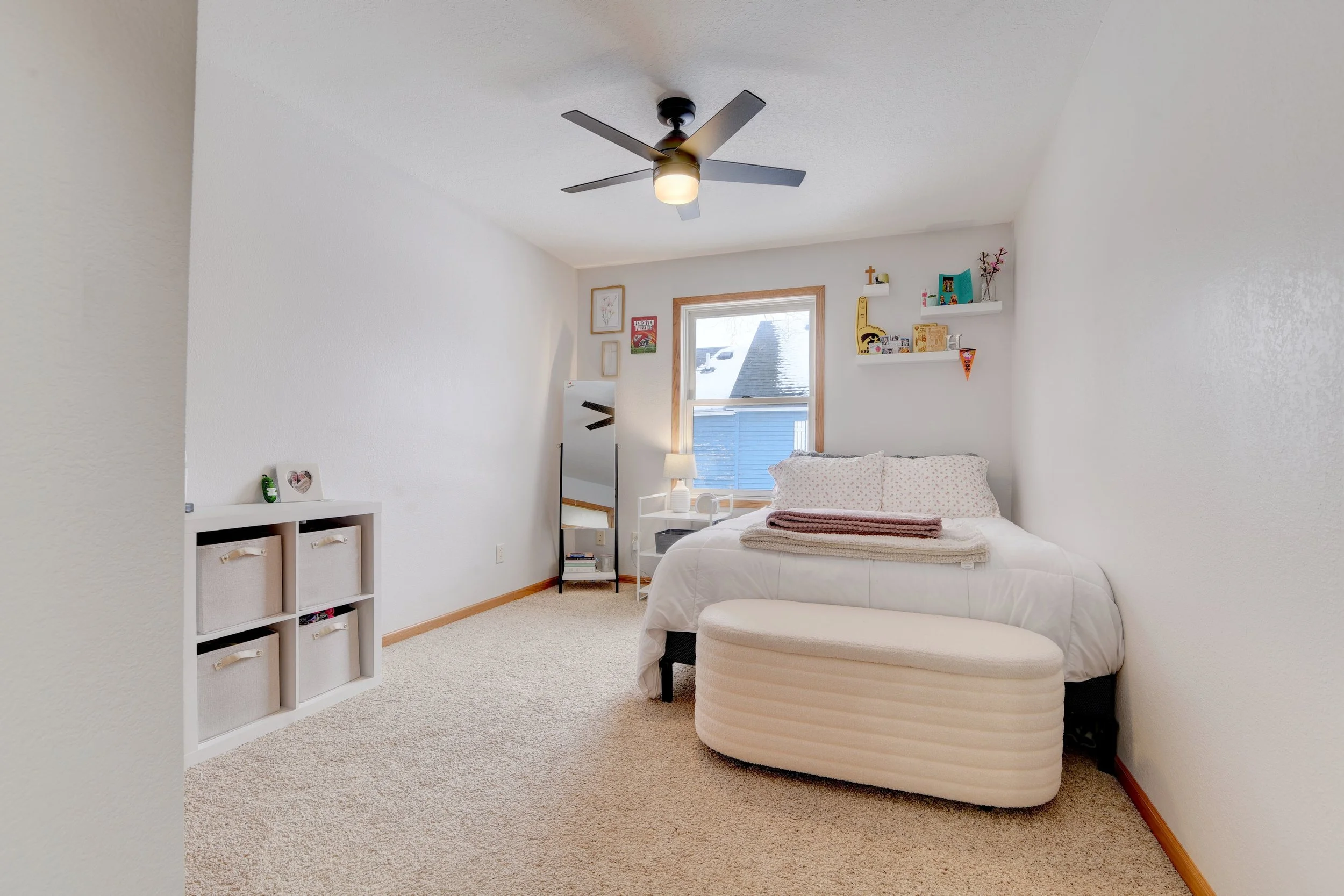 A bedroom with a ceiling fan, a window showing a snowy roof outside, a bed with folded blankets, a white storage bin, a mirror, a small white nightstand with a lamp, and floating shelves with decorations.