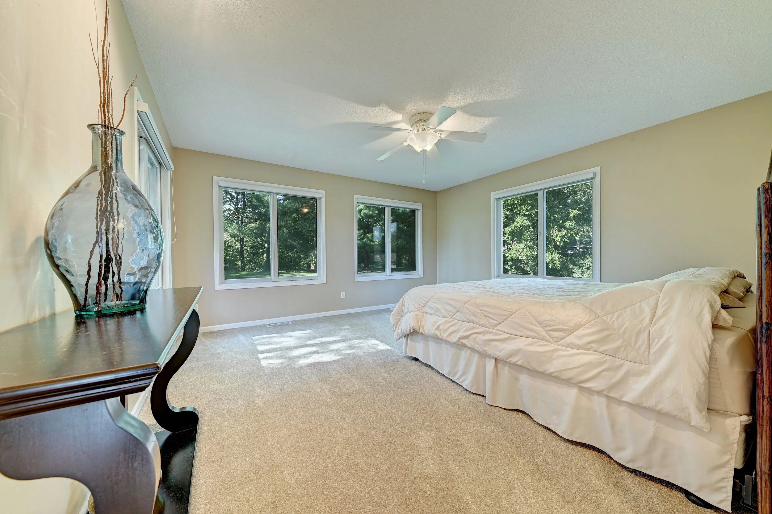 A bedroom with beige walls, a ceiling fan, large windows showing green trees outside, a bed with a white quilt, and a glass vase with decorative sticks on a wooden dresser.