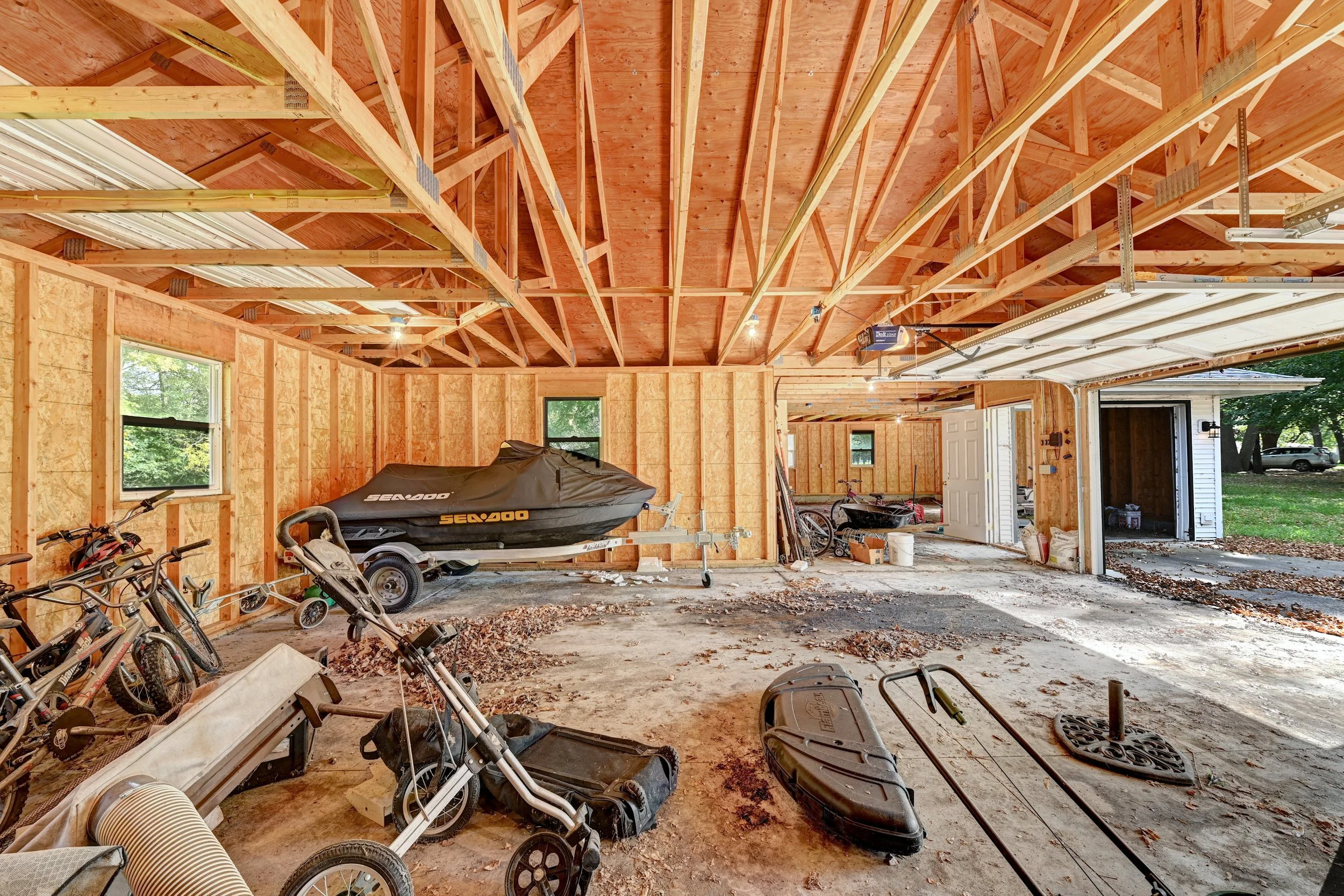 Interior of a house under construction with exposed wooden framing, including ceiling trusses and wall studs. Several bicycles, a boat trailer, and miscellaneous items are scattered on the unfinished concrete floor. The garage door is open, revealing