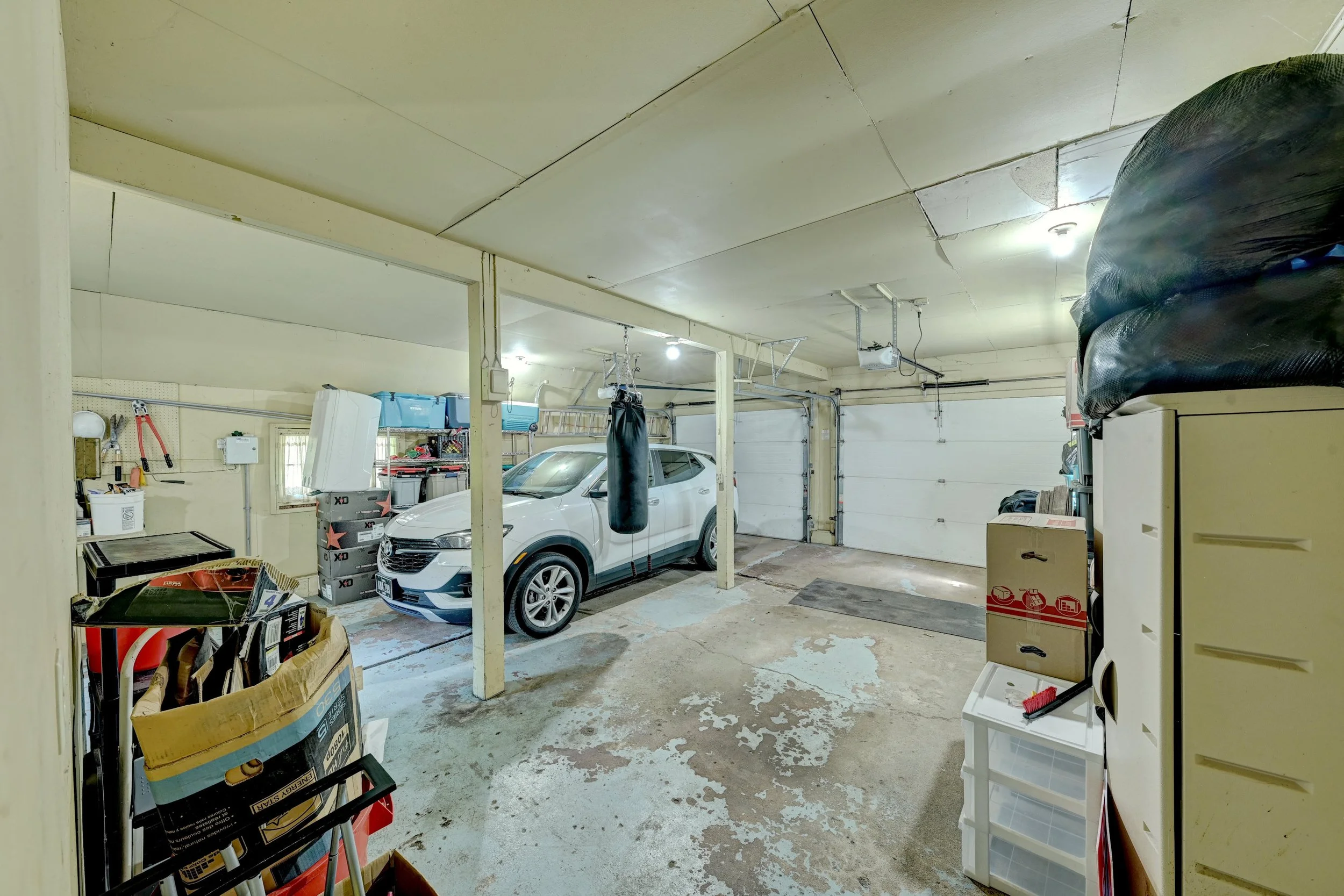 This image shows a spacious garage with a white vehicle parked inside. There are various storage items, boxes, and tools around the garage, with some hanging from the wall and others on shelves. The garage has a concrete floor, and the ceiling has vi