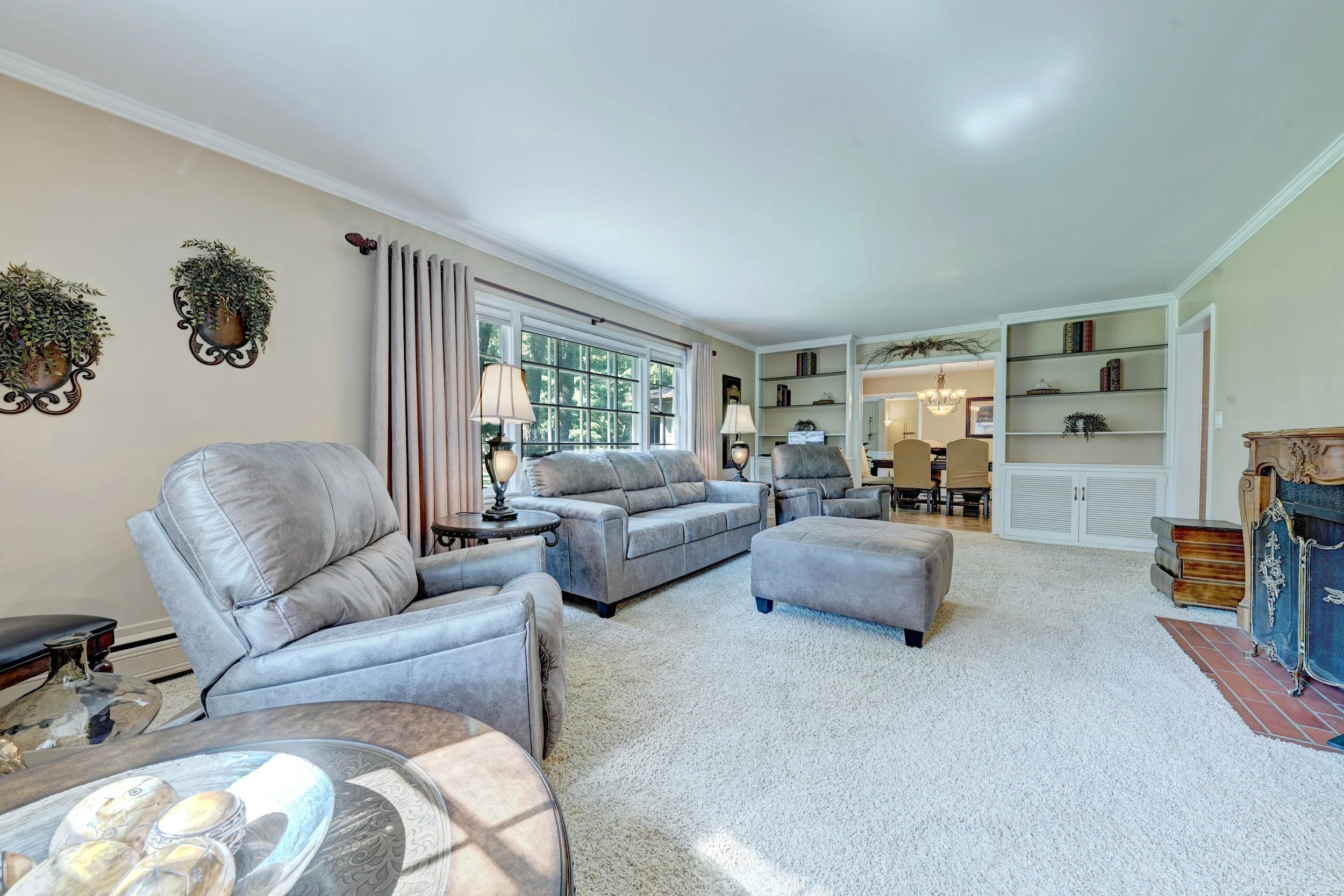 Living room with beige walls, large window with curtains, and built-in bookshelves. Contains gray furniture including a sofa, chairs, and a matching ottoman, with side tables and lamps. Part of a dining area with chairs and a chandelier visible in th