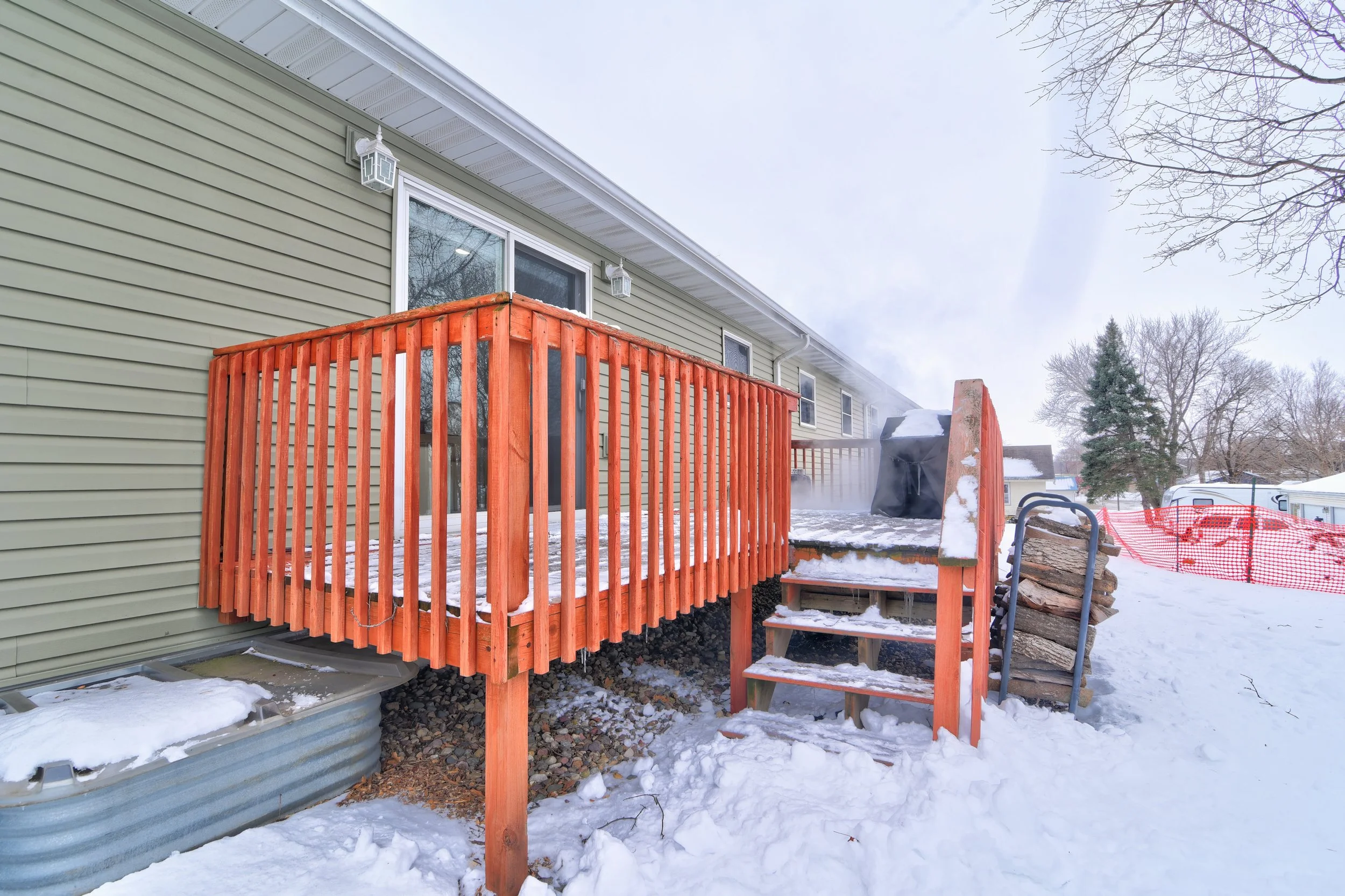 Snow-covered backyard with small wooden deck attached to house, stacked firewood, and trees in the background.