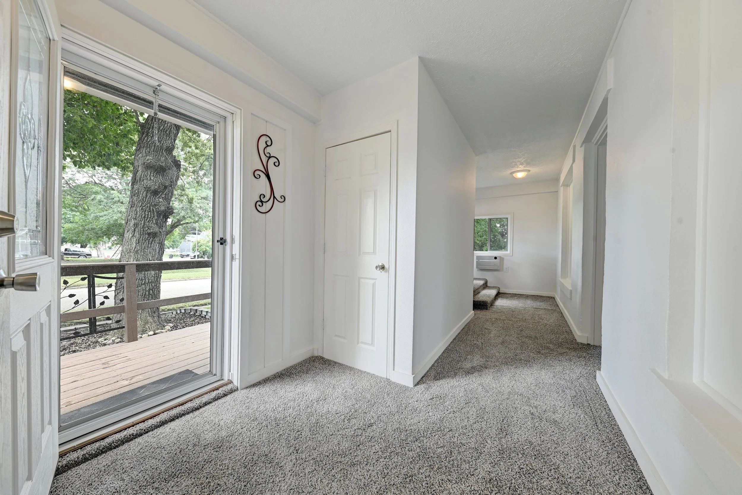 Interior view of a house entryway with a glass door leading to a small balcony or porch. The area has white painted walls, gray carpeting, and a staircase with carpeted steps. Outside, a large tree and parked cars are visible.