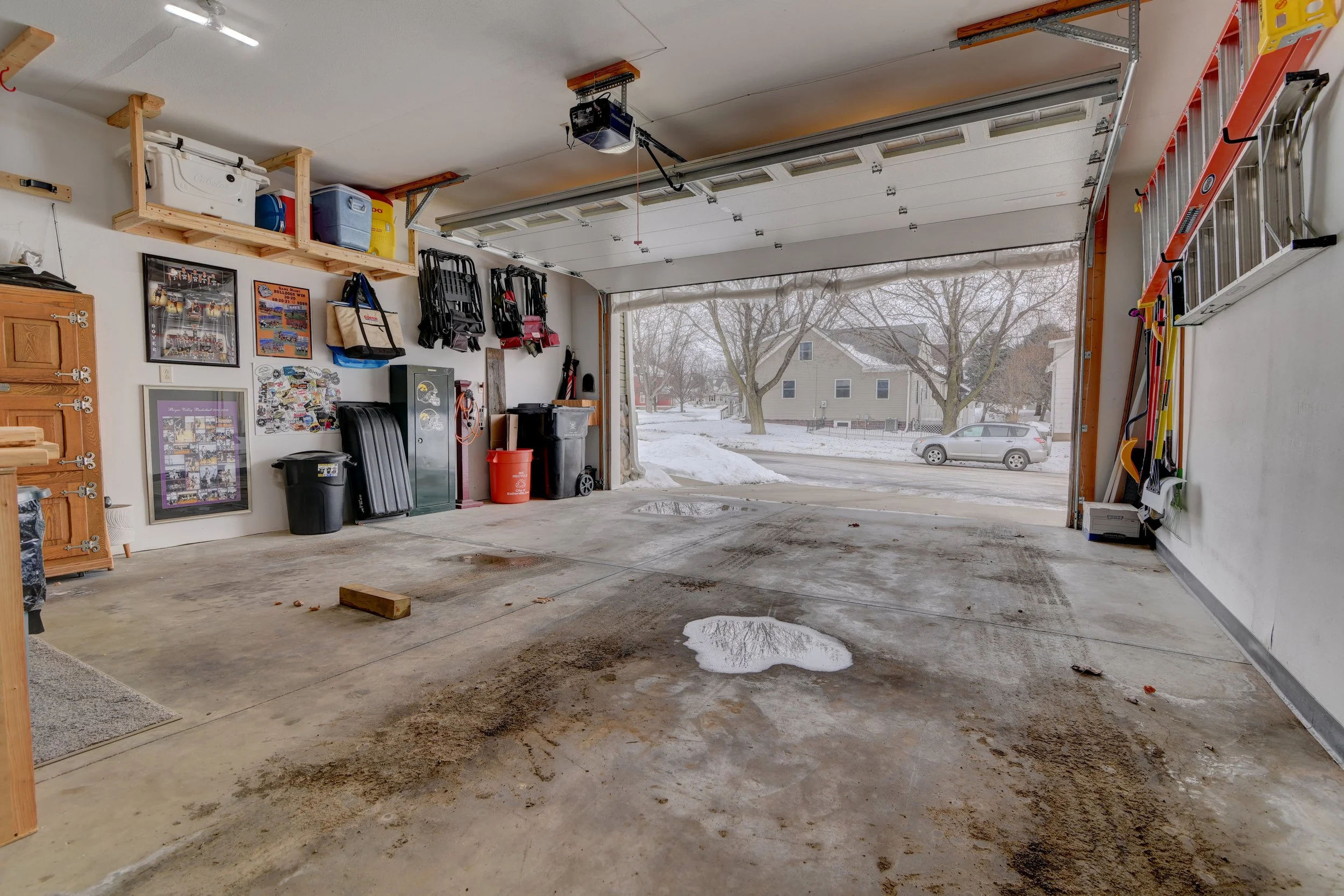 Empty garage with some water and dirt stains on the concrete floor, a few small puddles, and various tools and storage items organized along the walls. Outside is visible through the open garage door, with a snowy yard and a street with parked cars.