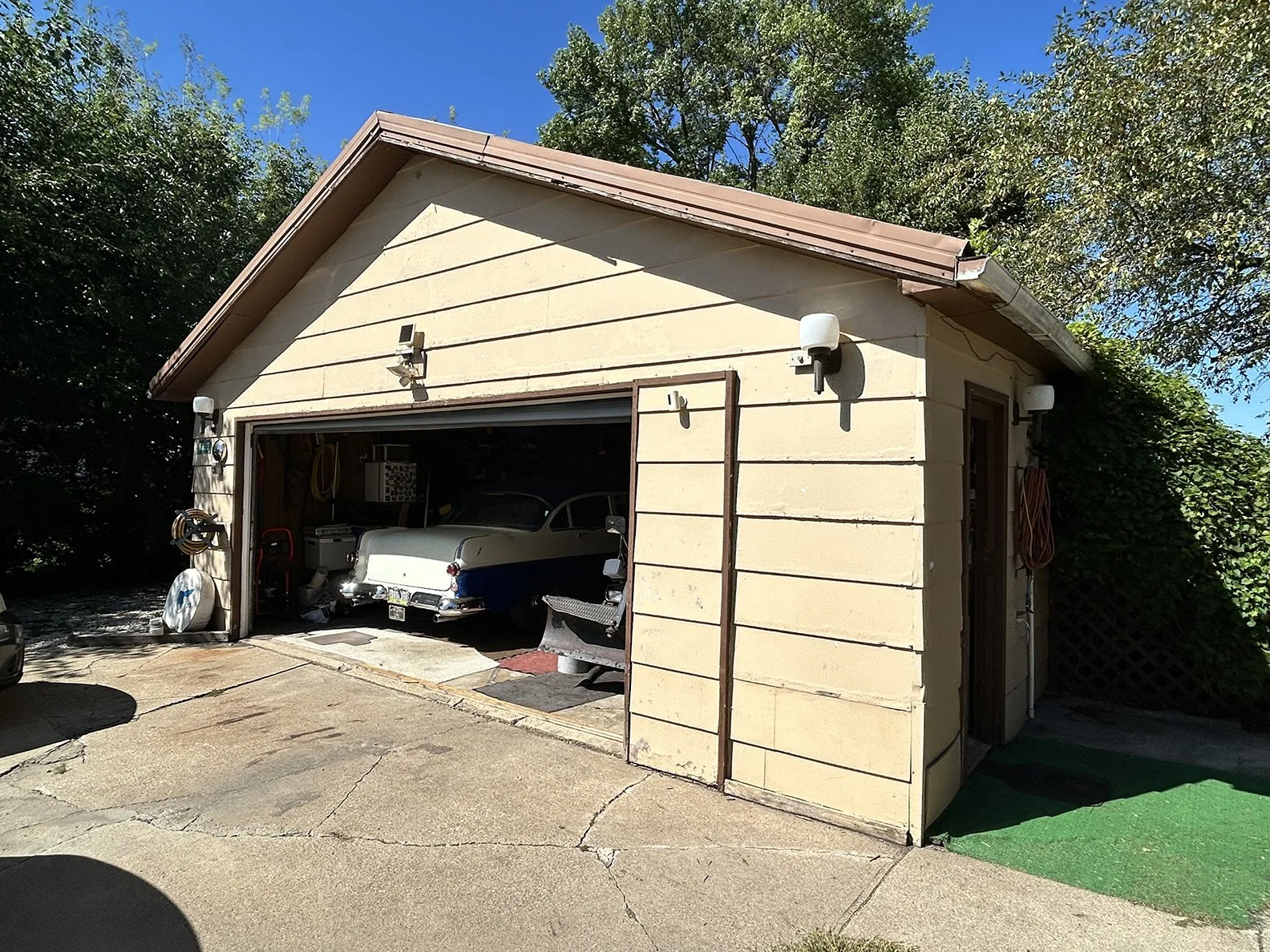 Garage with an open door containing a vintage car, surrounded by green trees and blue sky.