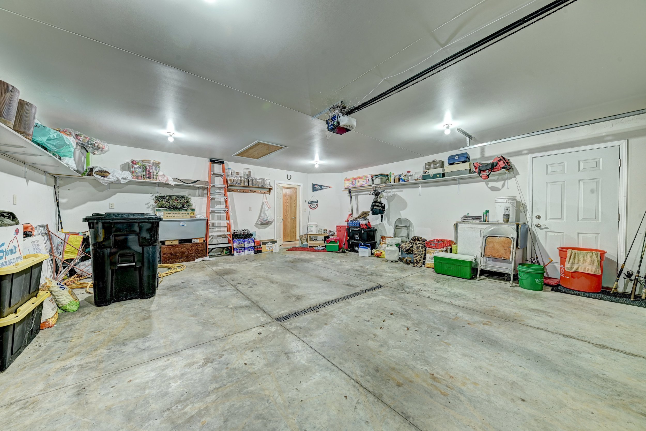 A cluttered garage with various storage shelves, tools, and containers, concrete floor, and a closed white door.