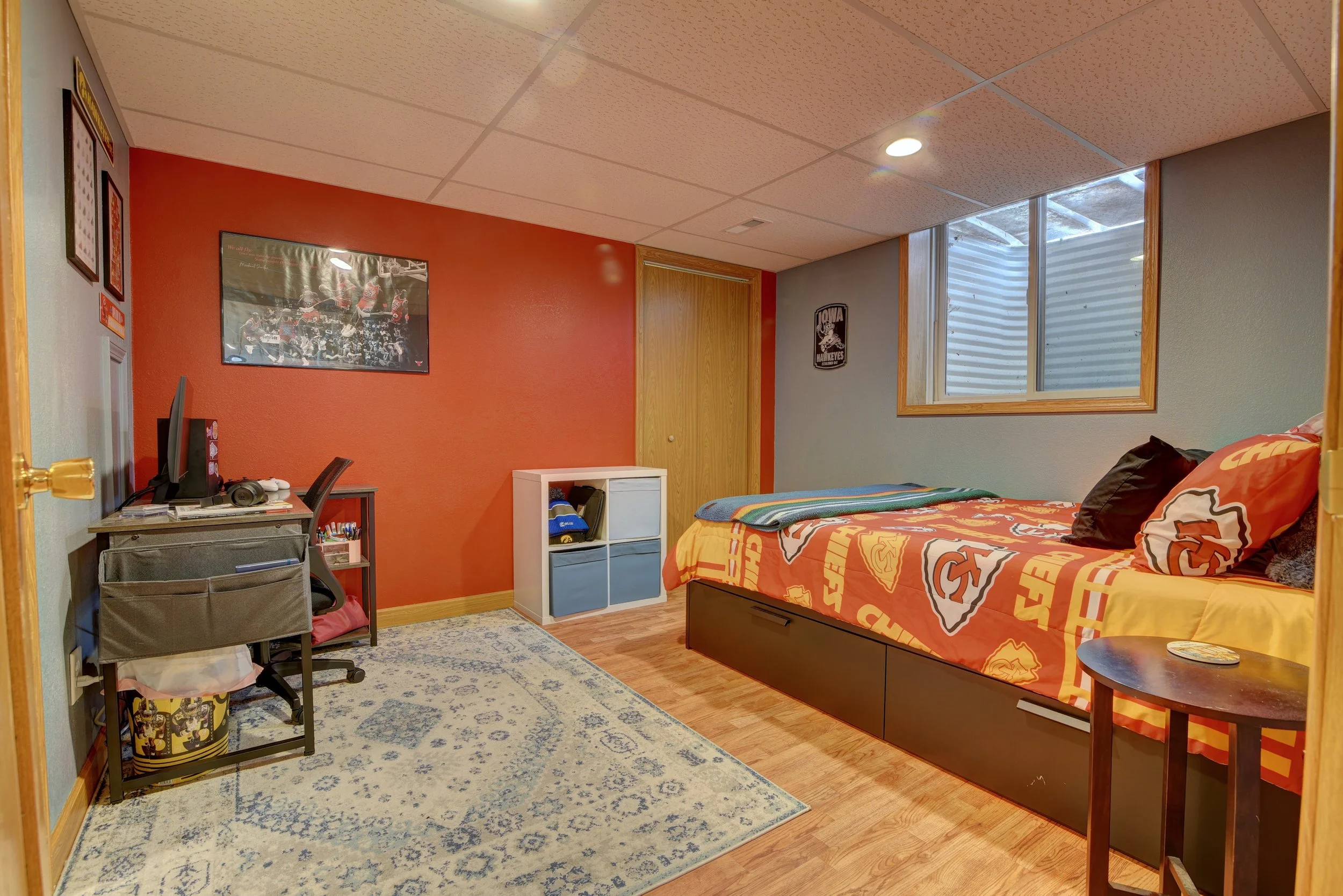 Bedroom with red accent wall, bed with sports-themed bedding, small side table, window with blinds, desk with computer, and framed sports posters.