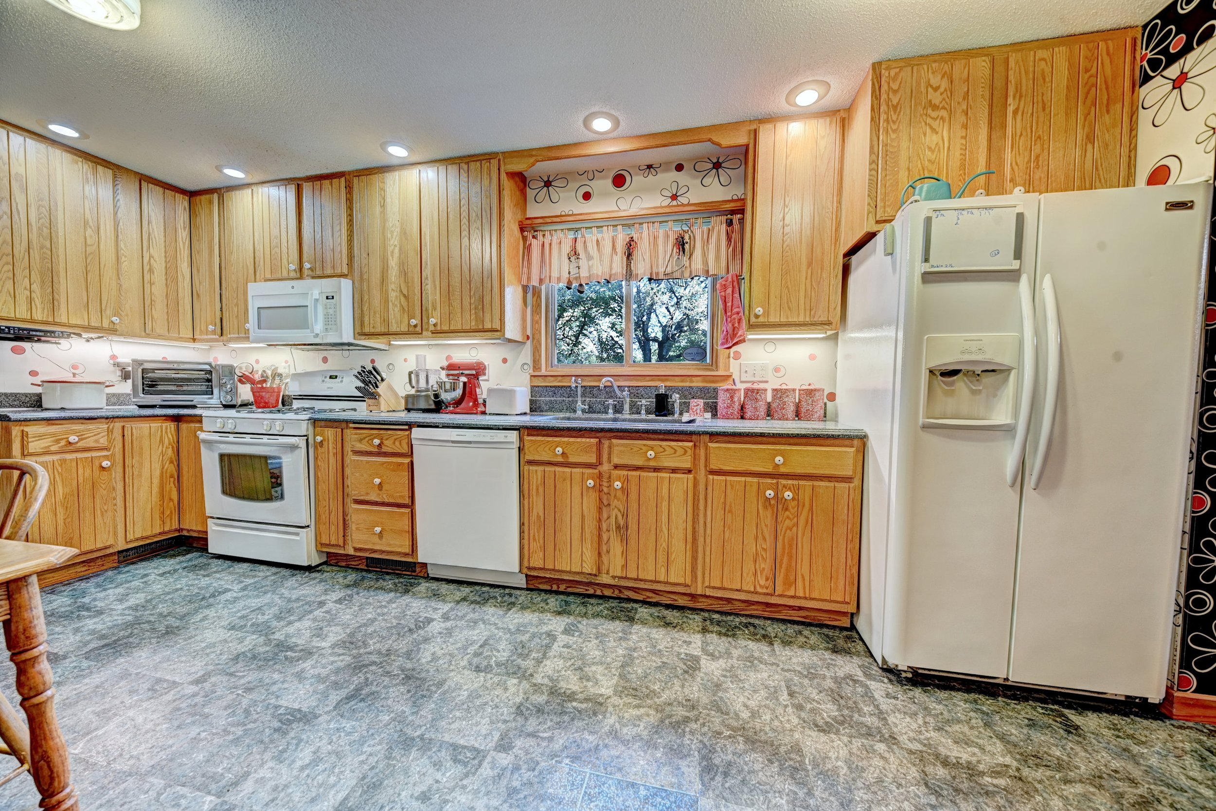 Kitchen with wooden cabinets, white appliances including a refrigerator and microwave, small window with pinkish curtains, and patterned wallpaper with red and black designs.