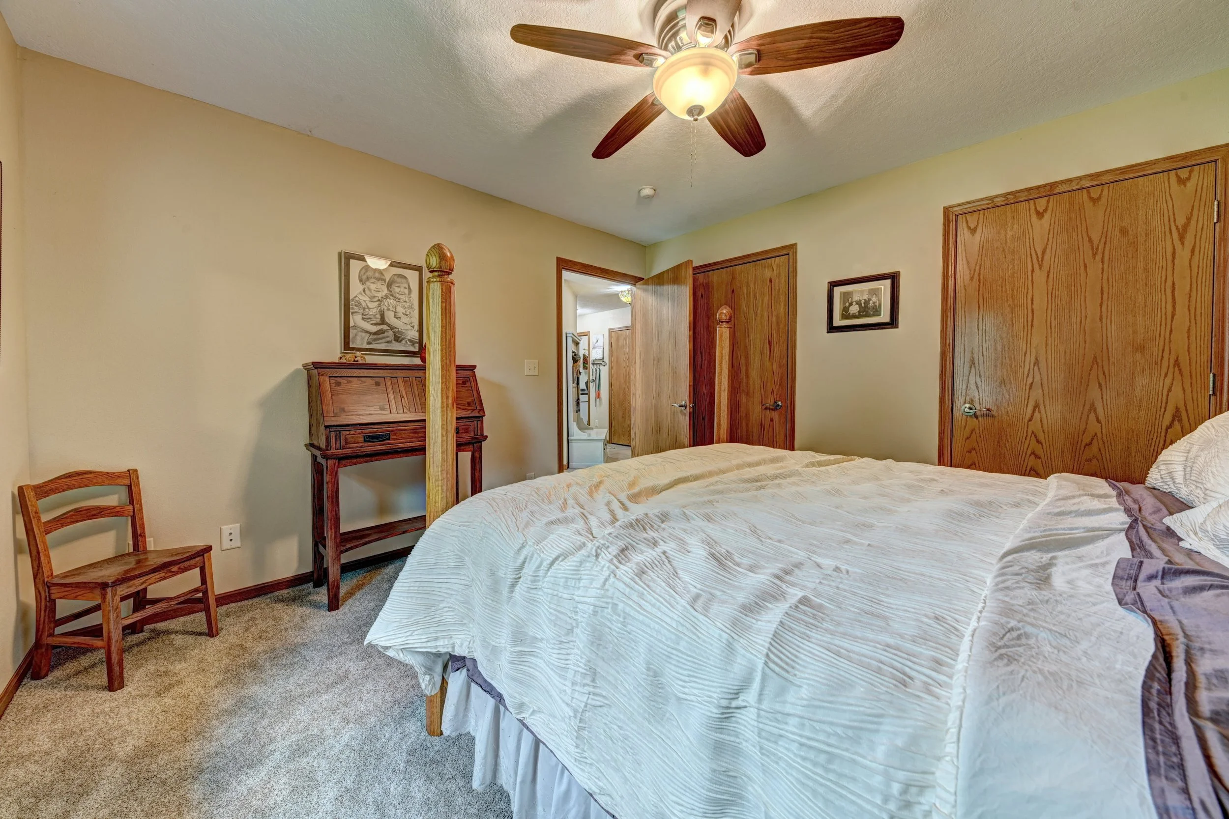 A bedroom with a ceiling fan, a bed with a cream-colored comforter, wooden doors, framed black and white photographs on the walls, a wooden writing desk, and a chair. Another room is visible through the open door.