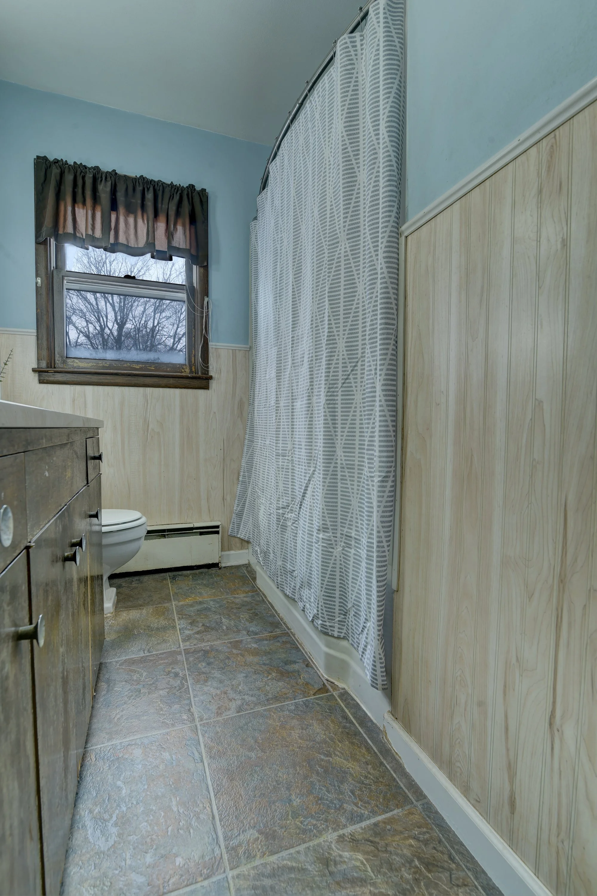 Interior of a small bathroom with a window, toilet, vanity, and a shower with a patterned curtain.