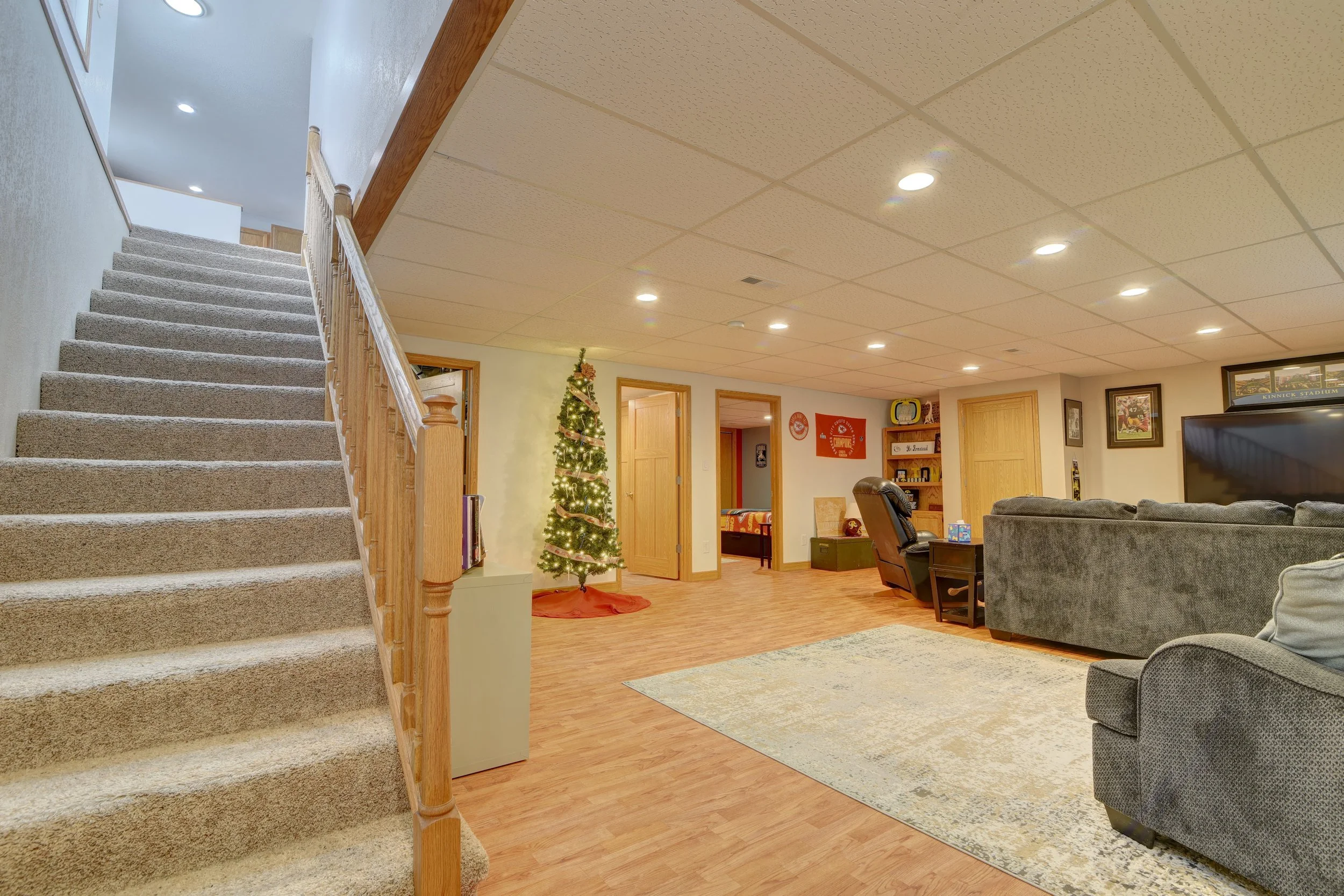 Living room with a staircase on the left, decorated Christmas tree, beige carpets, grey sofas, television, and wooden doors on the wall.