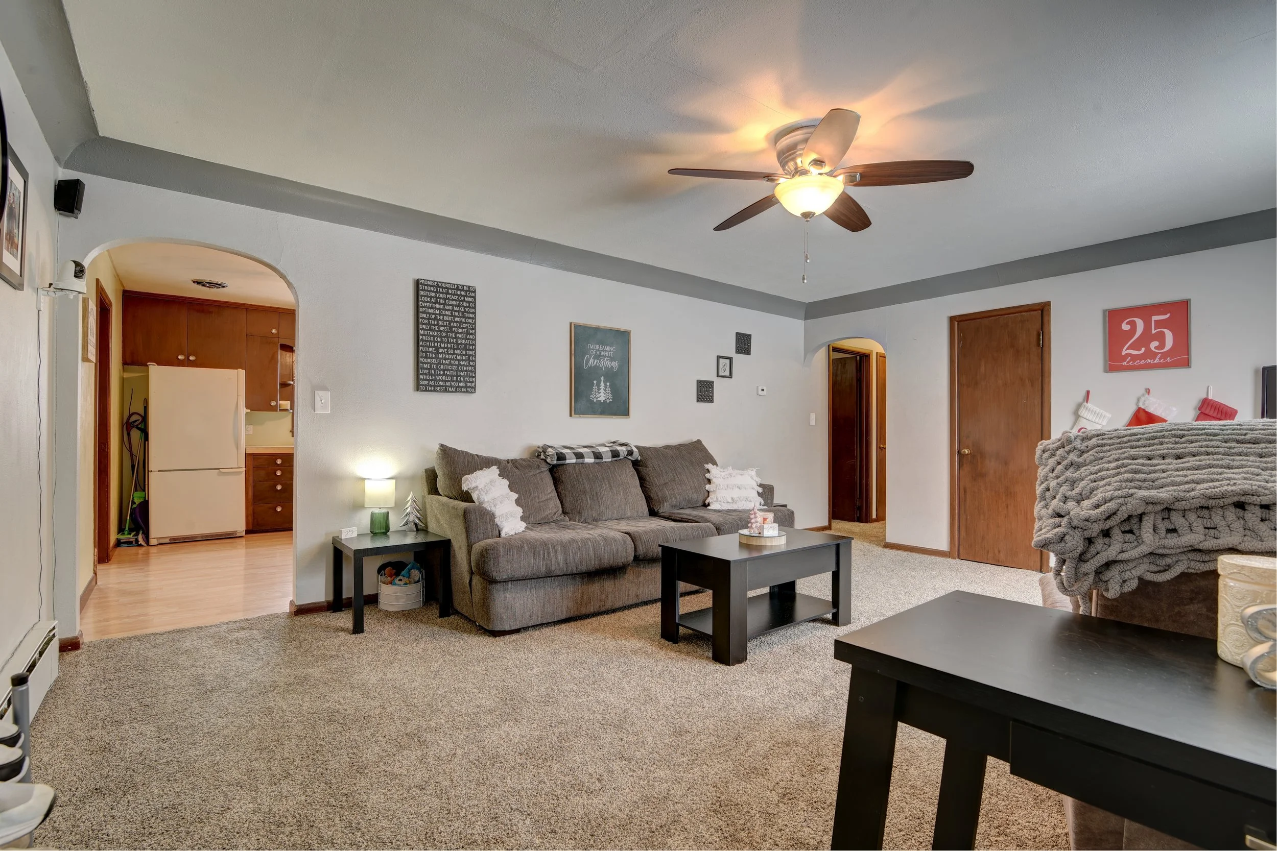 Living room with a brown sofa, black coffee table, and a ceiling fan. Kitchen with a refrigerator is visible through an arched doorway.