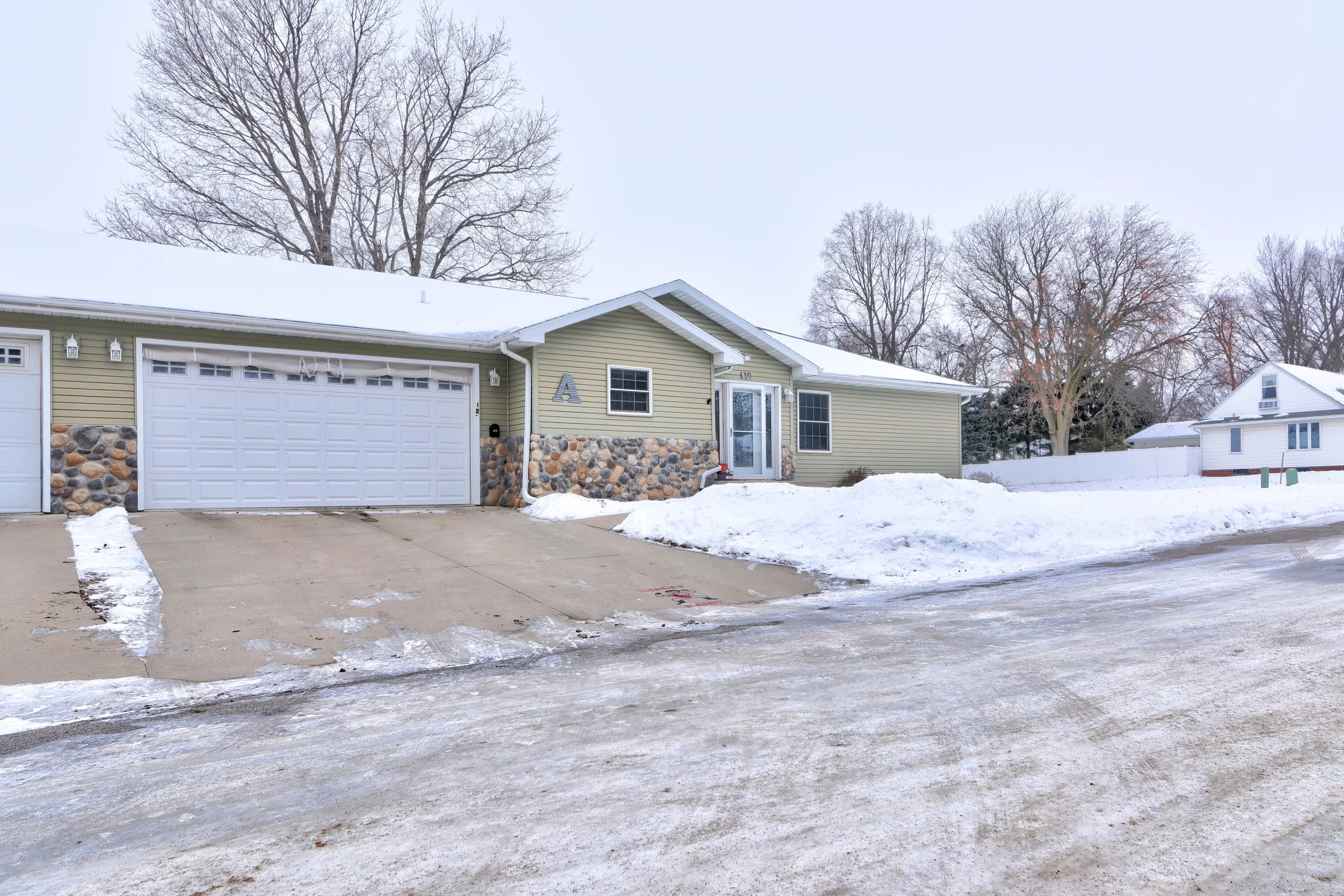 A single-story house with a green exterior, stone accents, and a white garage door, surrounded by snow and leafless trees in a residential neighborhood.