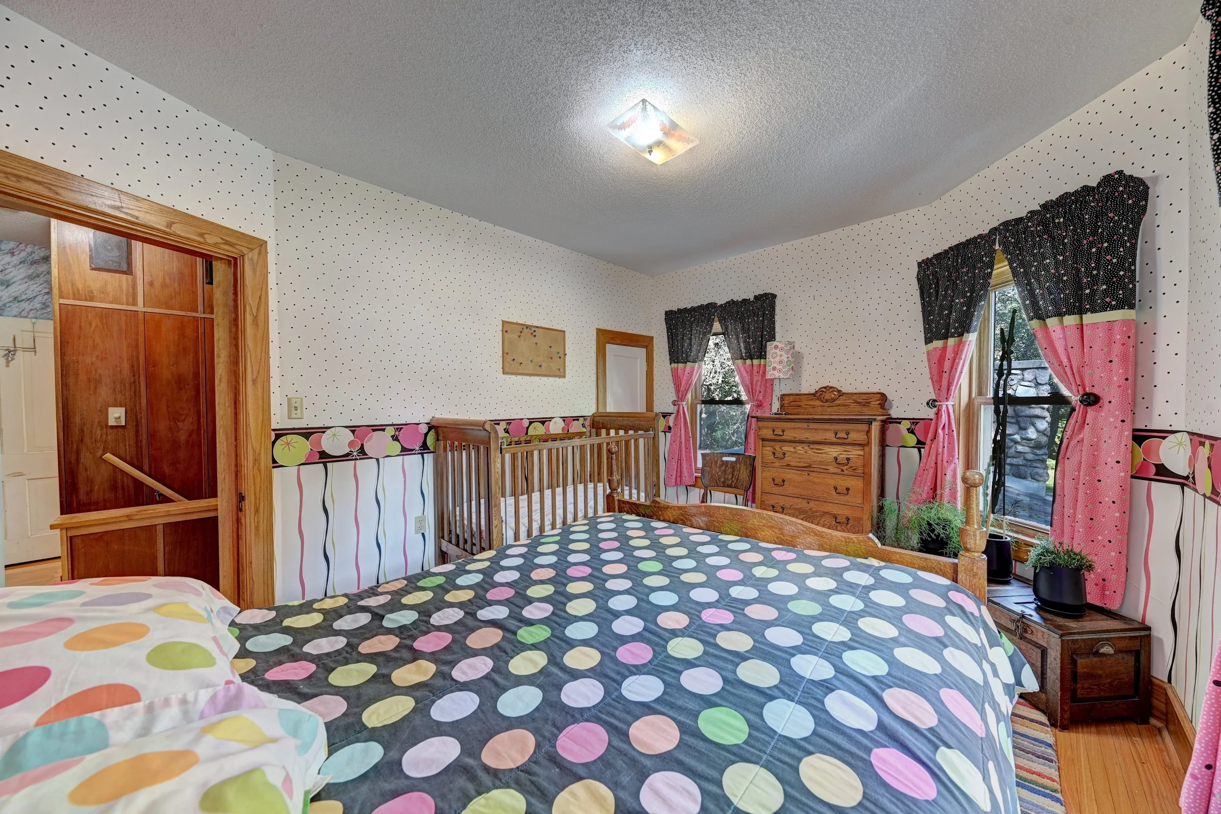 A bedroom with polka dot bedding and curtains featuring pink and black colors, wood furniture, and windows showing a stone wall outside.