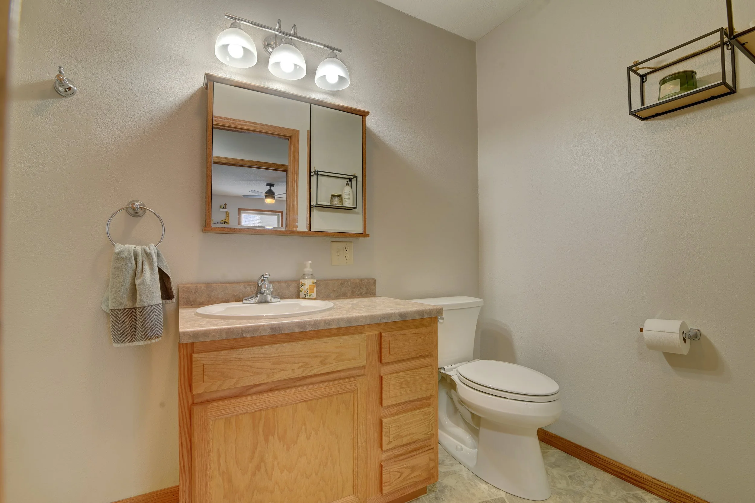 A small bathroom with a wooden vanity, a sink, and a mirror cabinet. There is a beige countertop, a soap dispenser, and a hand towel hanging on a wall-mounted towel ring. To the right is a toilet with a toilet paper roll attached to the wall, and abo