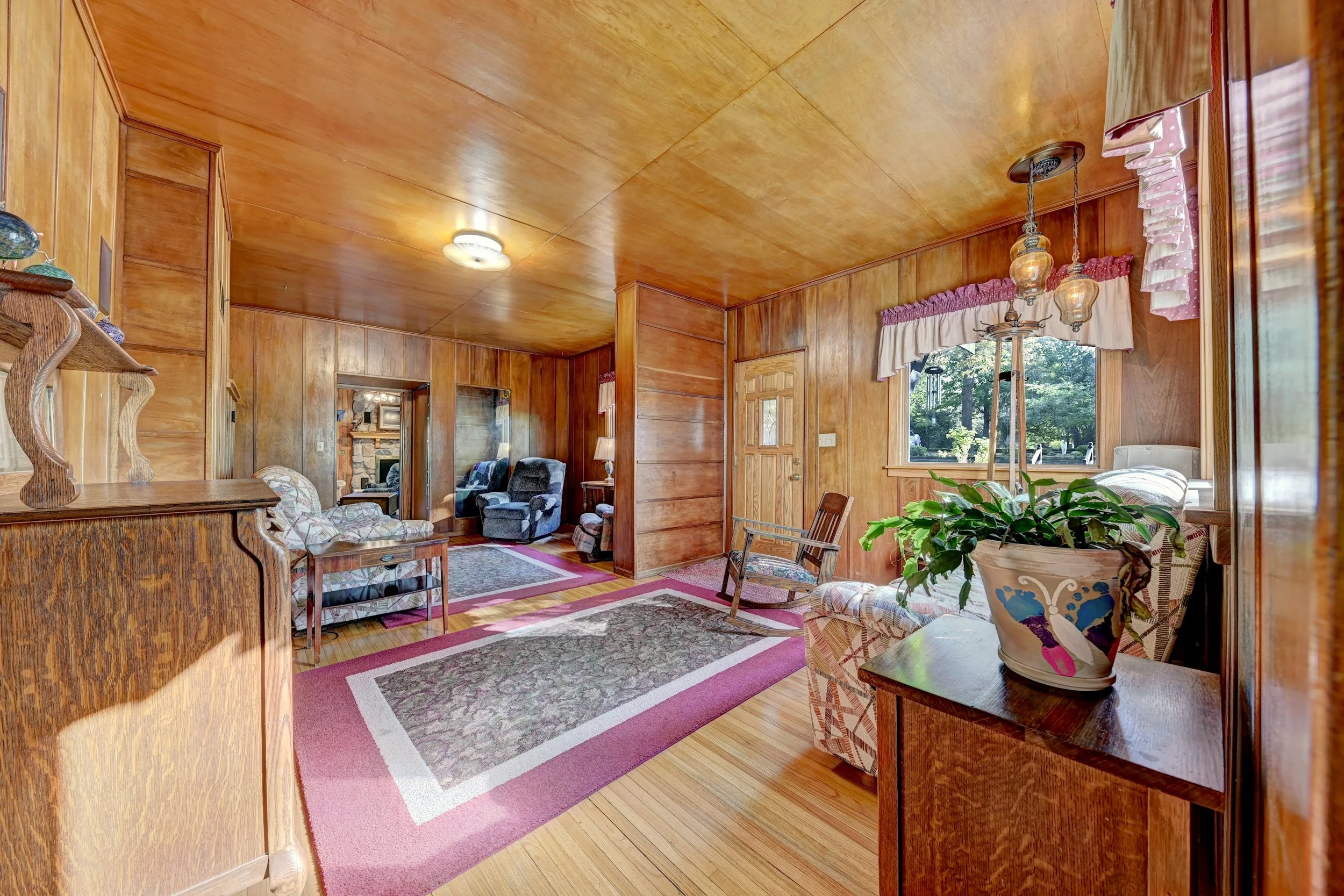 Living room with wood-paneled walls and ceiling, furnished with armchairs, a floral and patterned chair, a wooden coffee table, curtains with pink ruffles, and a potted plant near a large window with scenic outdoor view.