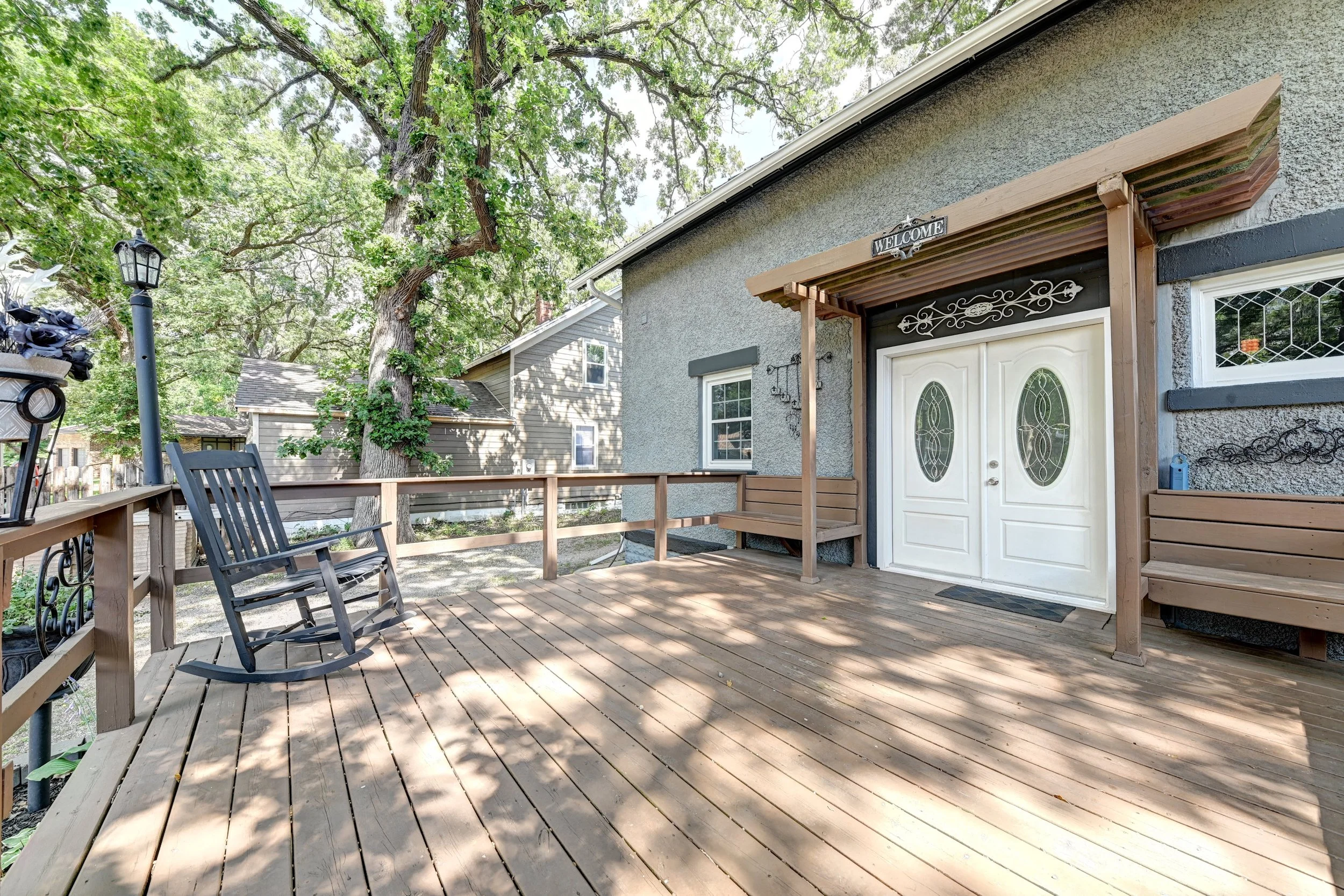 A backyard deck with a wooden railing and benches, surrounded by lush green trees, a house with a textured gray exterior, and a decorative lamp post.
