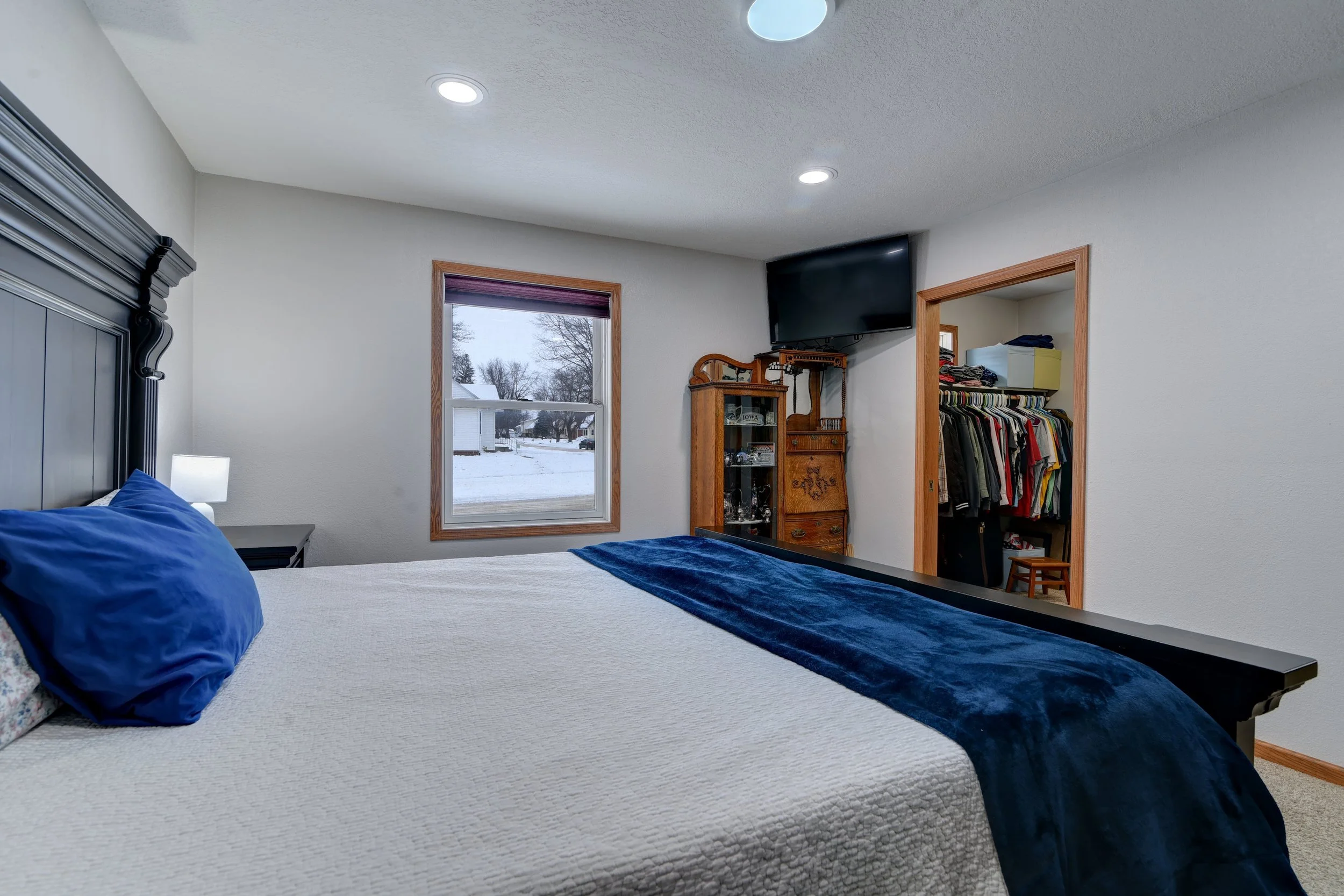 A bedroom with a large bed covered with a beige quilt, blue pillows, and a blue velvet throw. There is a window with a view of a snowy yard, a nightstand with a lamp, and a closet with clothes inside. A small TV is mounted on the wall above an antiqu