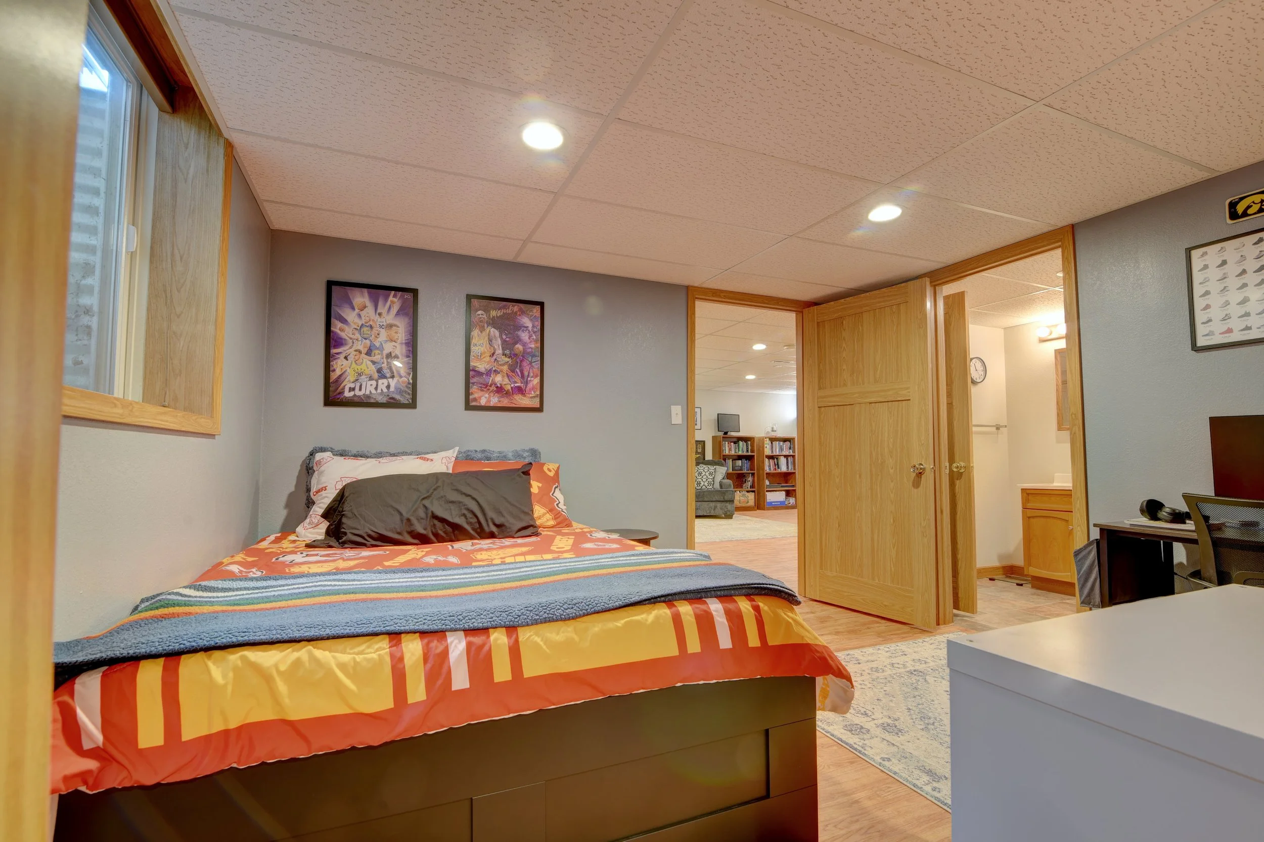 A bedroom with a bed covered in colorful orange, yellow, and blue bedding, two sports posters on the wall, a window to the left, and an open doorway leading to a living room and a bathroom.