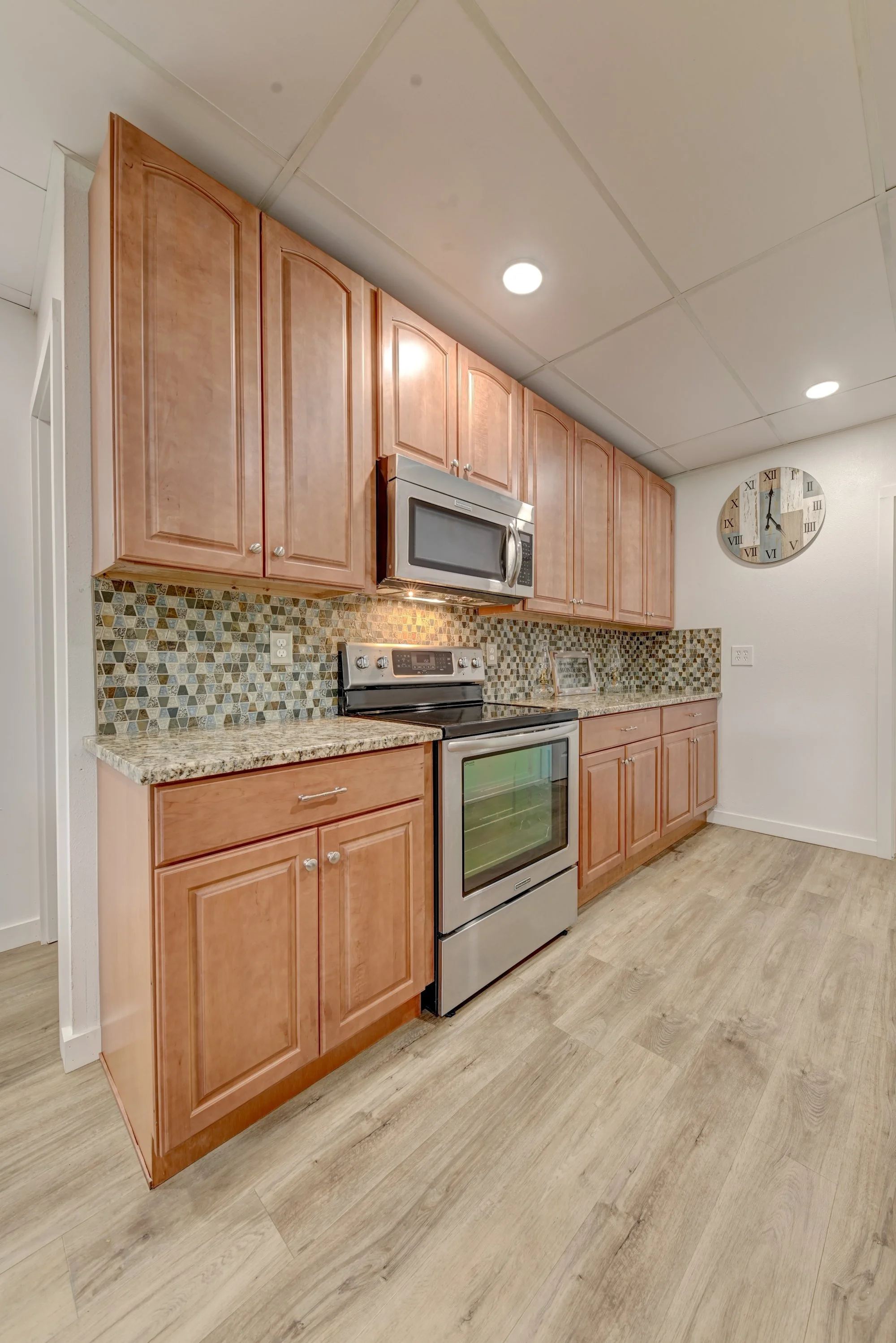 Kitchen with wooden cabinets, granite countertops, a stainless steel microwave and oven, a mosaic tile backsplash, a wall clock, and wood flooring.