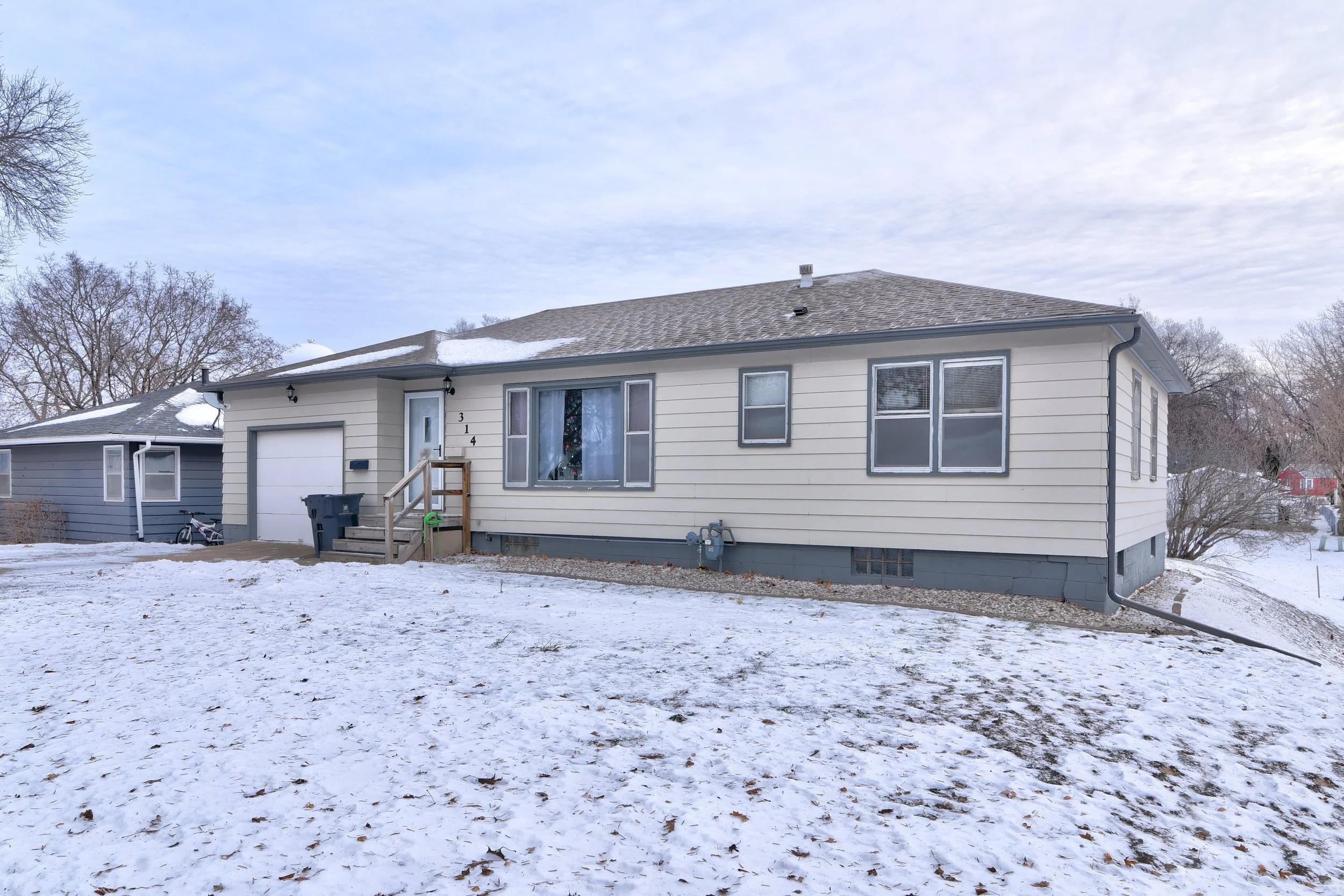 A house with white siding and a gray roof, surrounded by snow, with a small front porch and a garage on the left side.