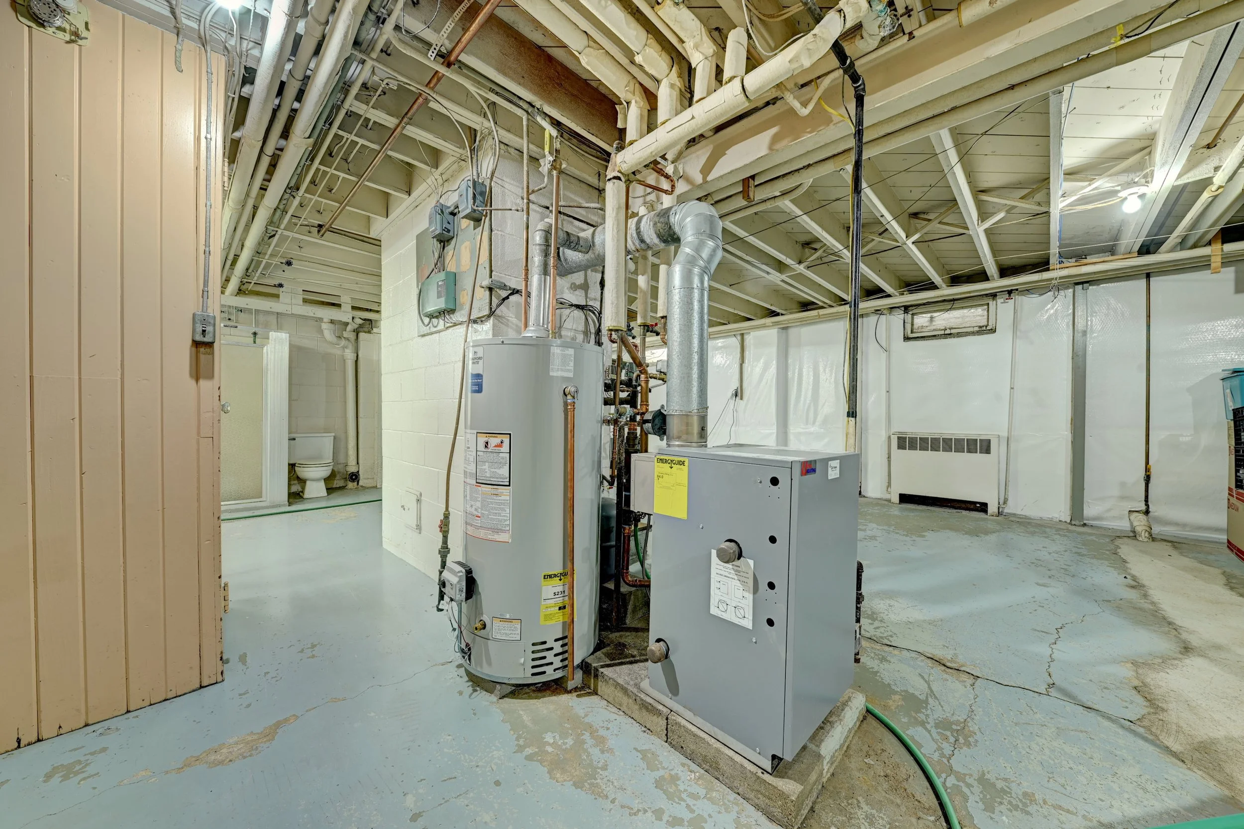 Interior of a basement or utility room with a water heater, furnace, exposed pipes, and a small toilet in the background.
