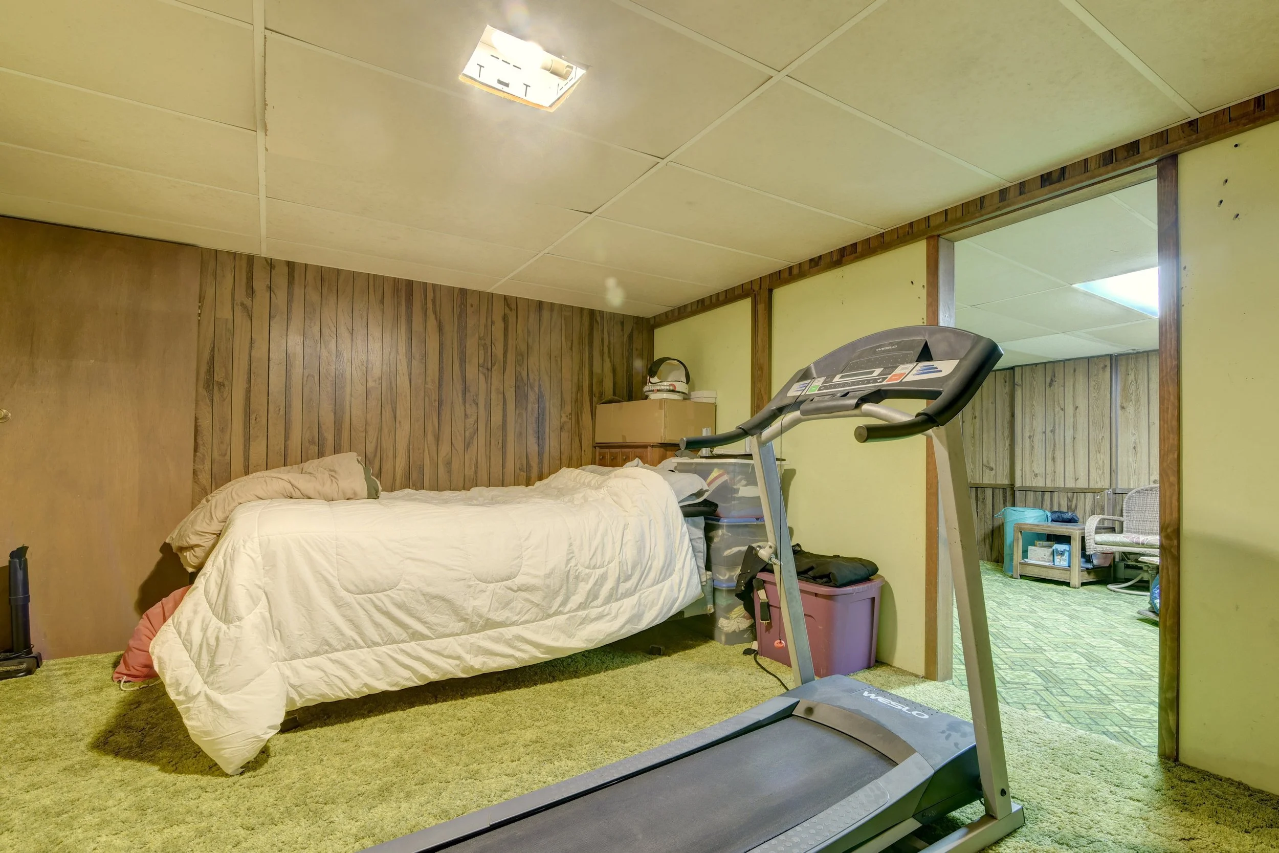 A small basement room with wood-paneled walls, a bed with a beige comforter and pillow, a treadmill, and a separate sitting area with a chair and table.
