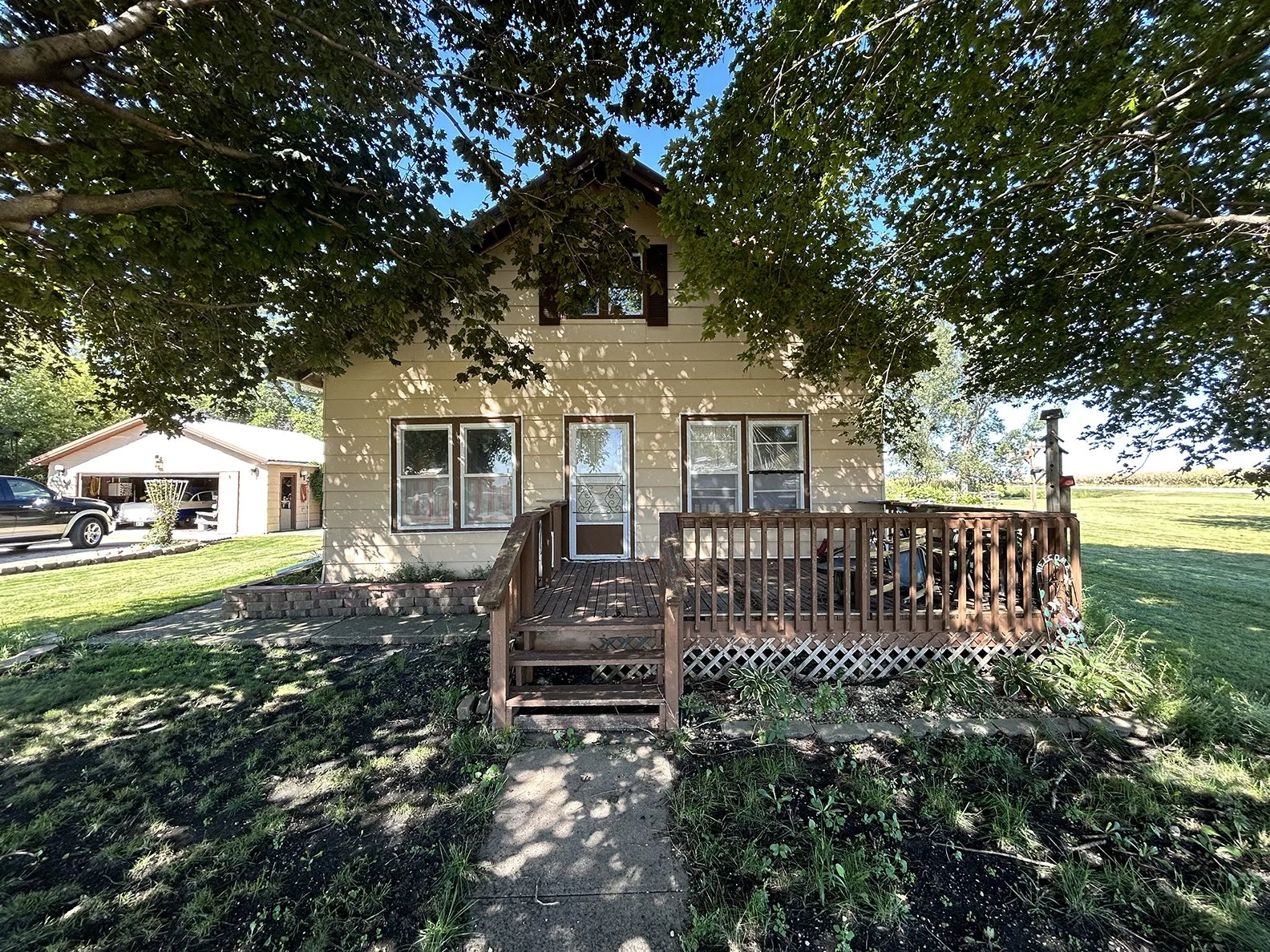 Back view of a yellow house with a wooden deck, multiple windows, and a porch door, surrounded by greenery and a large tree with sprawling branches.