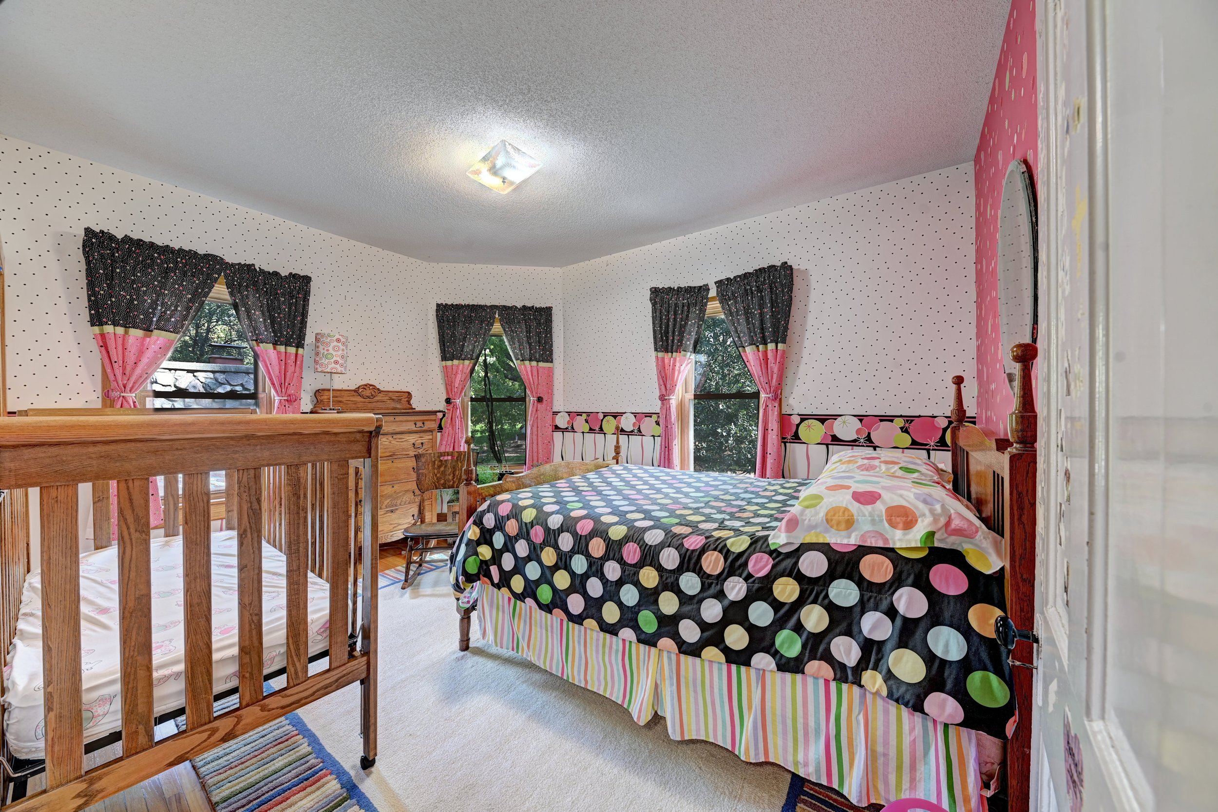A bedroom with polka dot wallpaper, pink and black curtains, a wooden bed frame with a polka dot bedspread, and a crib with a floral sheet. There is a ceiling light fixture, a window with a view of trees, and a rainbow striped area rug.