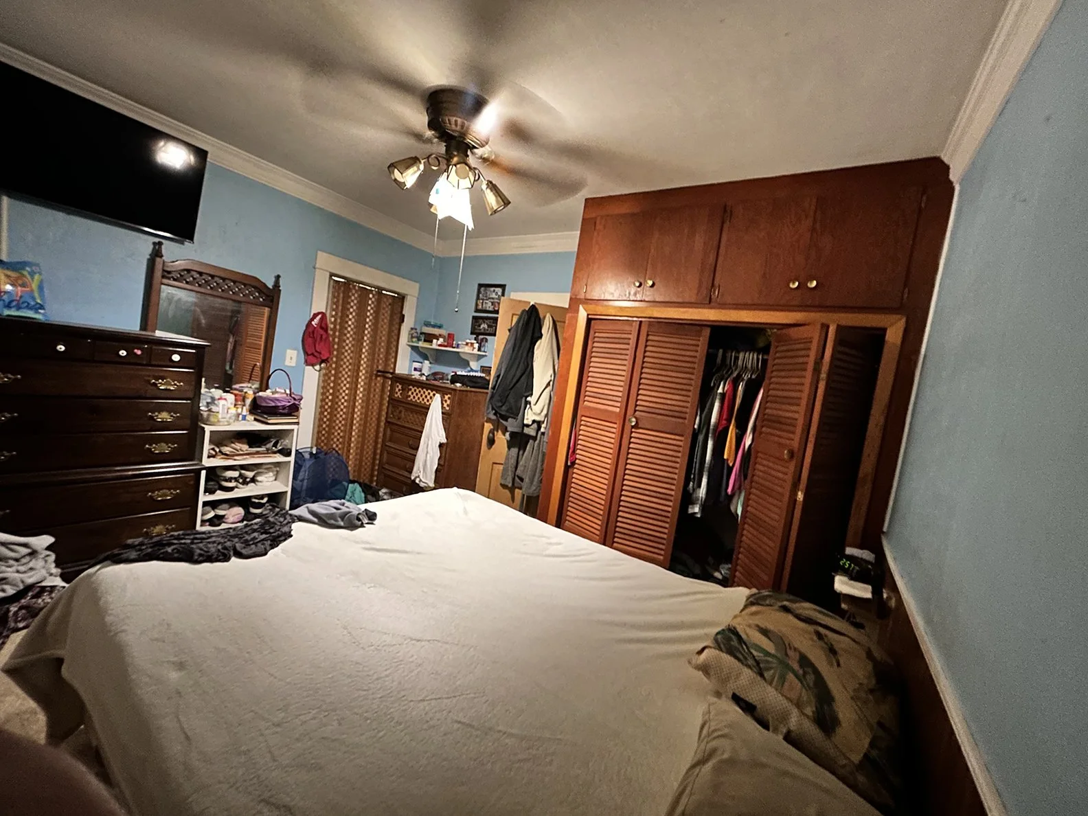This image shows a bedroom with blue walls, a dark wood dresser, a white shoe rack, a bed with a beige cover, and an open closet with clothing inside. There is a ceiling fan with lights, a flat-screen TV mounted on the wall, and various clutter like 