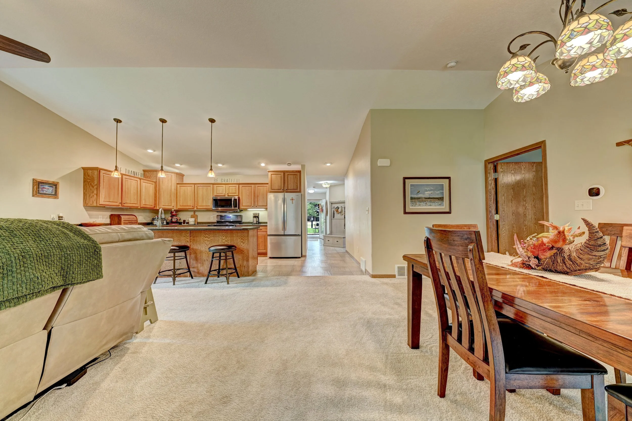 Open-concept living room and kitchen with wooden cabinets, stainless steel appliances, dining table with chairs, and pendant and ceiling lights in a home interior.