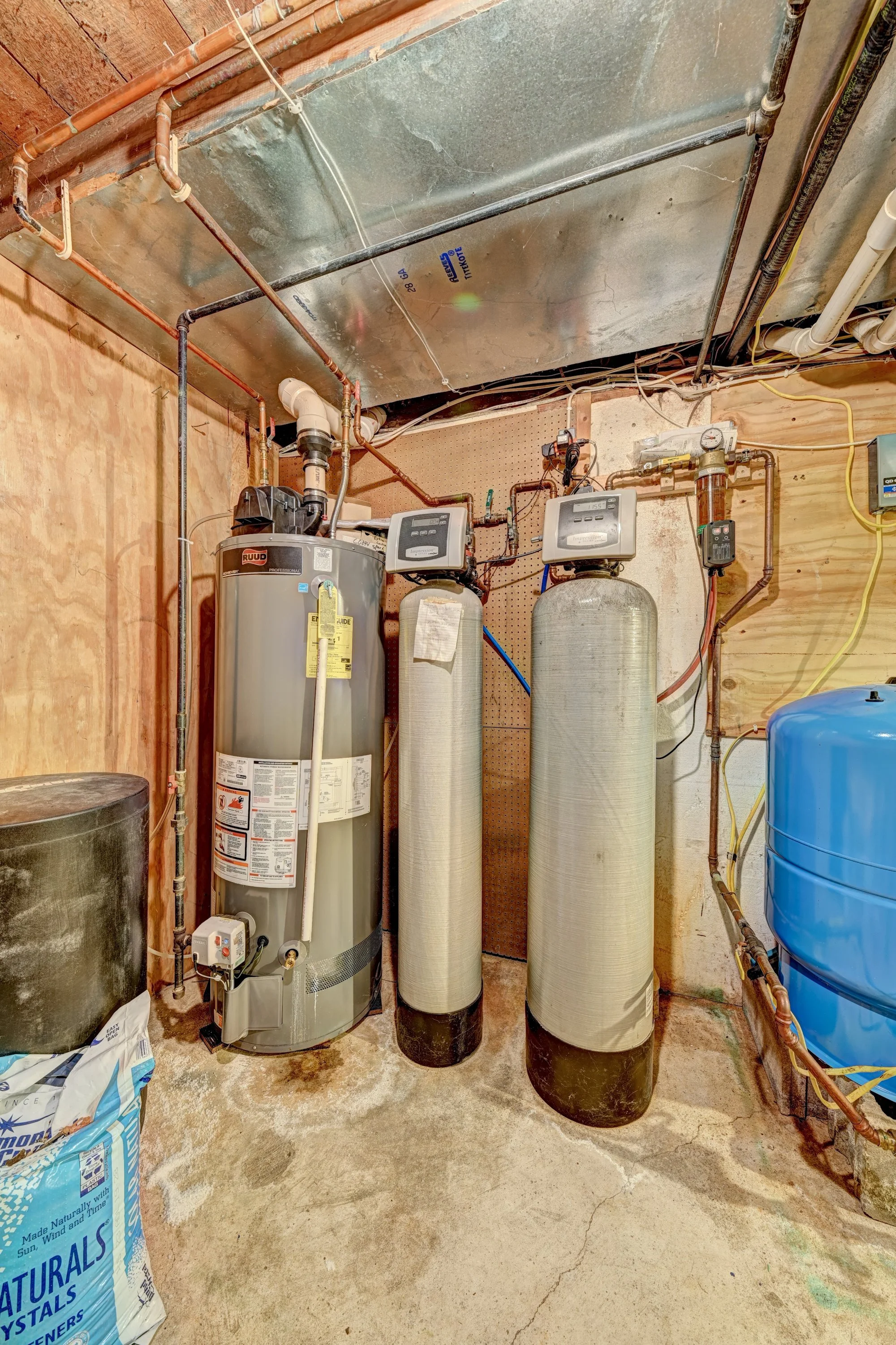 Room with water heater, water softener tanks, and plumbing pipes against a plywood wall and concrete floor.