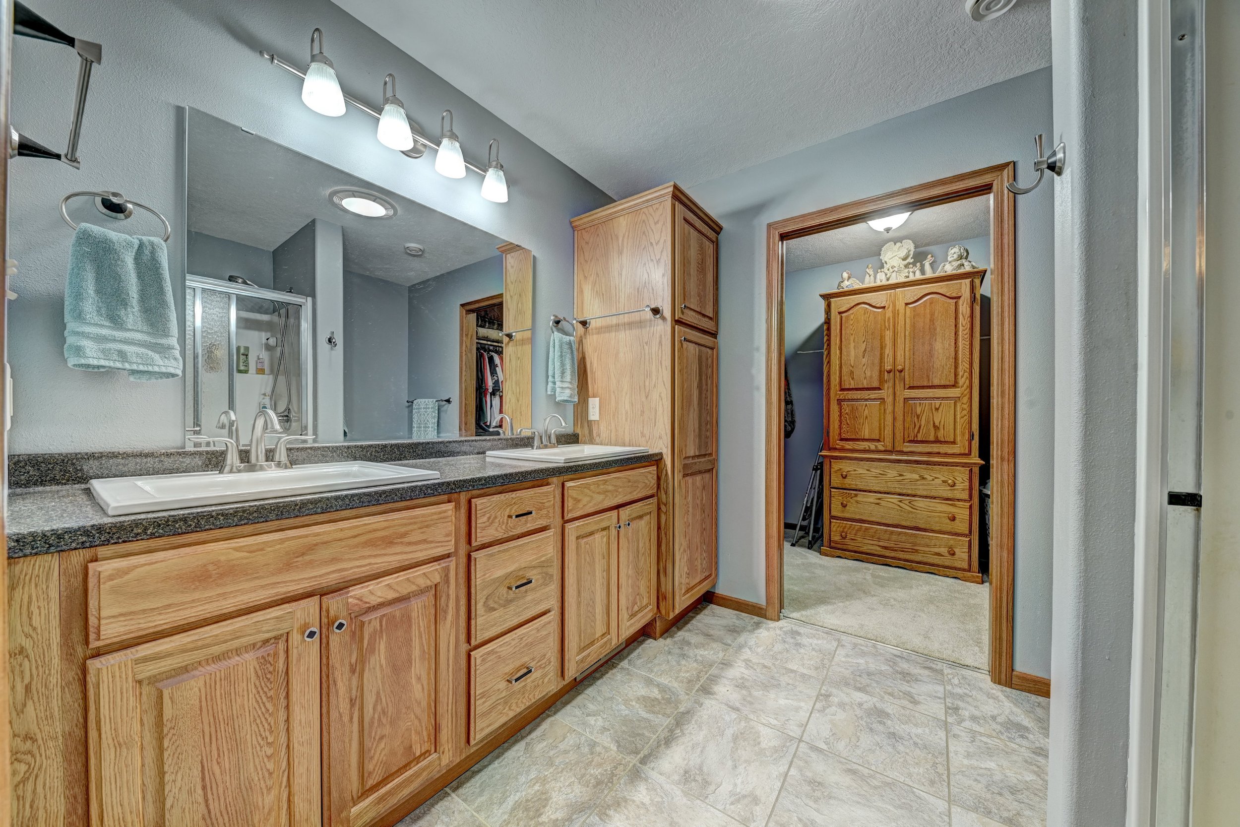 A bathroom with a double sink vanity with wooden cabinets and granite countertop, a large mirror, wooden medicine cabinet, wooden closet door, and tiled floor.