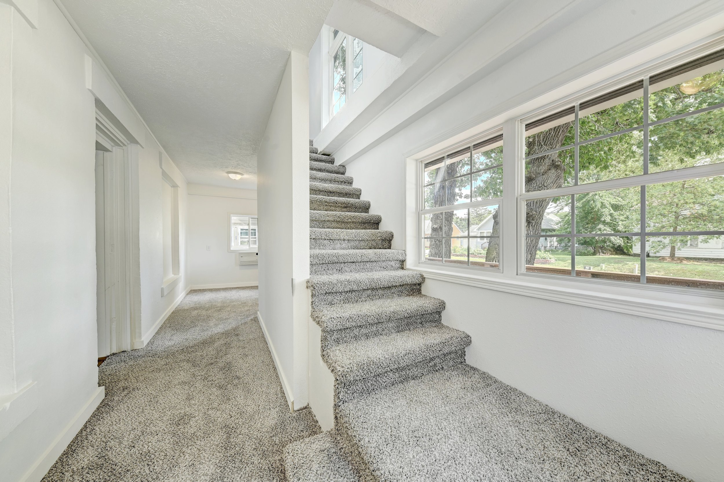 Interior view of a bright stairway with large windows, grey carpeted stairs, and white walls.
