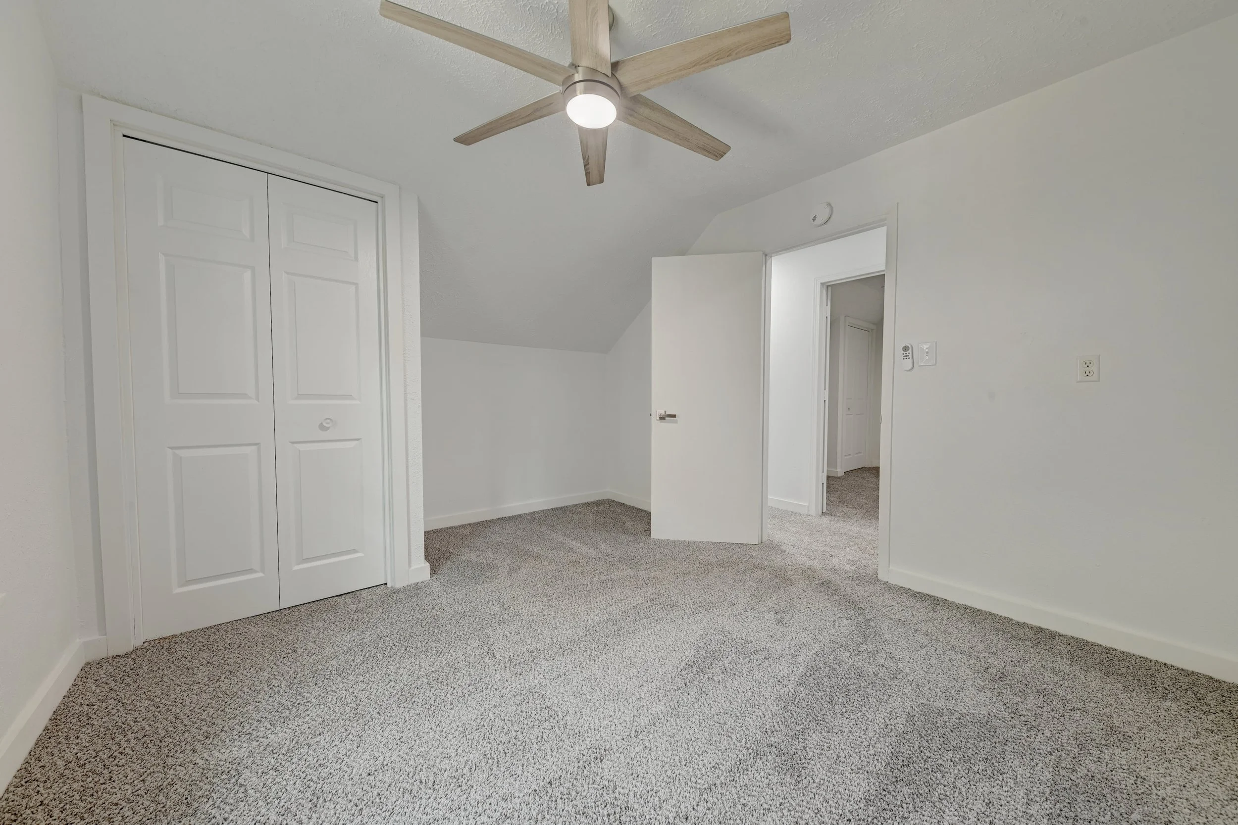 Empty room with gray carpet, white walls, a closet with double doors, an open door leading to another room, and a ceiling fan with wooden blades and a light inside.