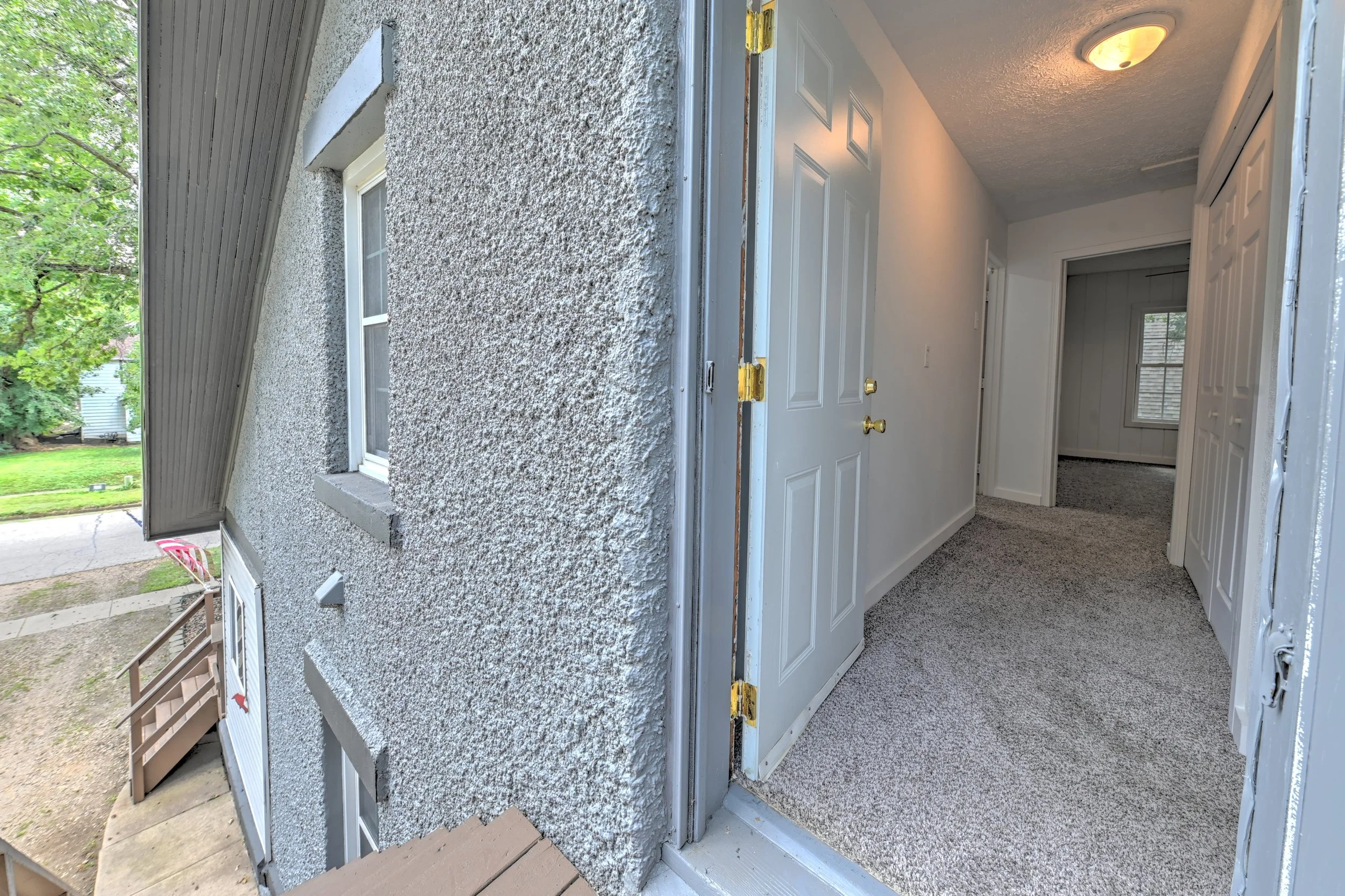 Photo of a house exterior with a view from the porch, showing textured stucco wall, a window, and a white door. The door is open, revealing a room with white walls and carpeted flooring. A ceiling light fixture is visible inside.