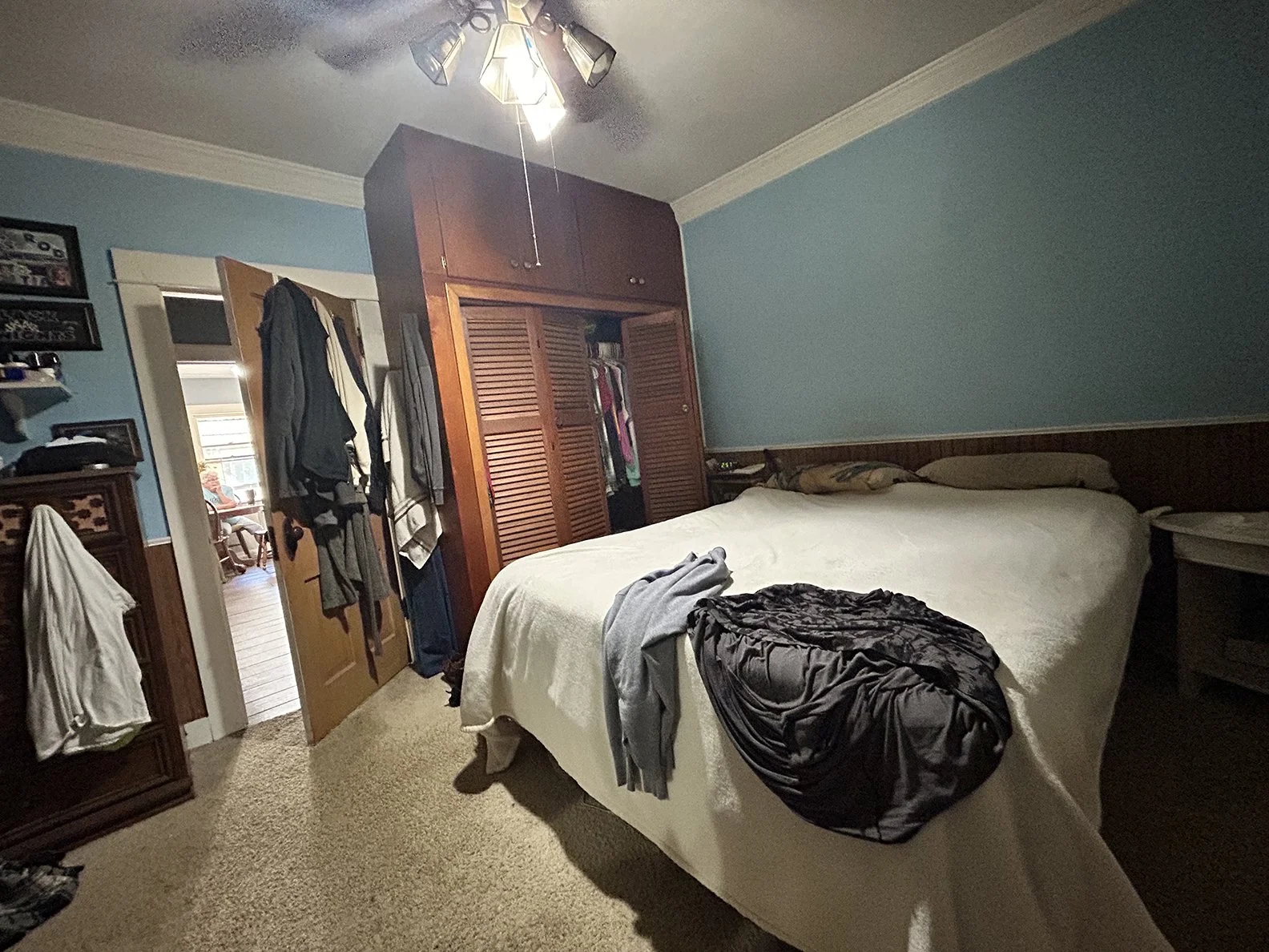 A bedroom with blue walls, a bed with a white bedspread and pillows, wooden closet doors with clothes hanging inside, a ceiling fan with lights, and a doorway leading to a separate room with a chair and window. Clothes are draped over furniture and o