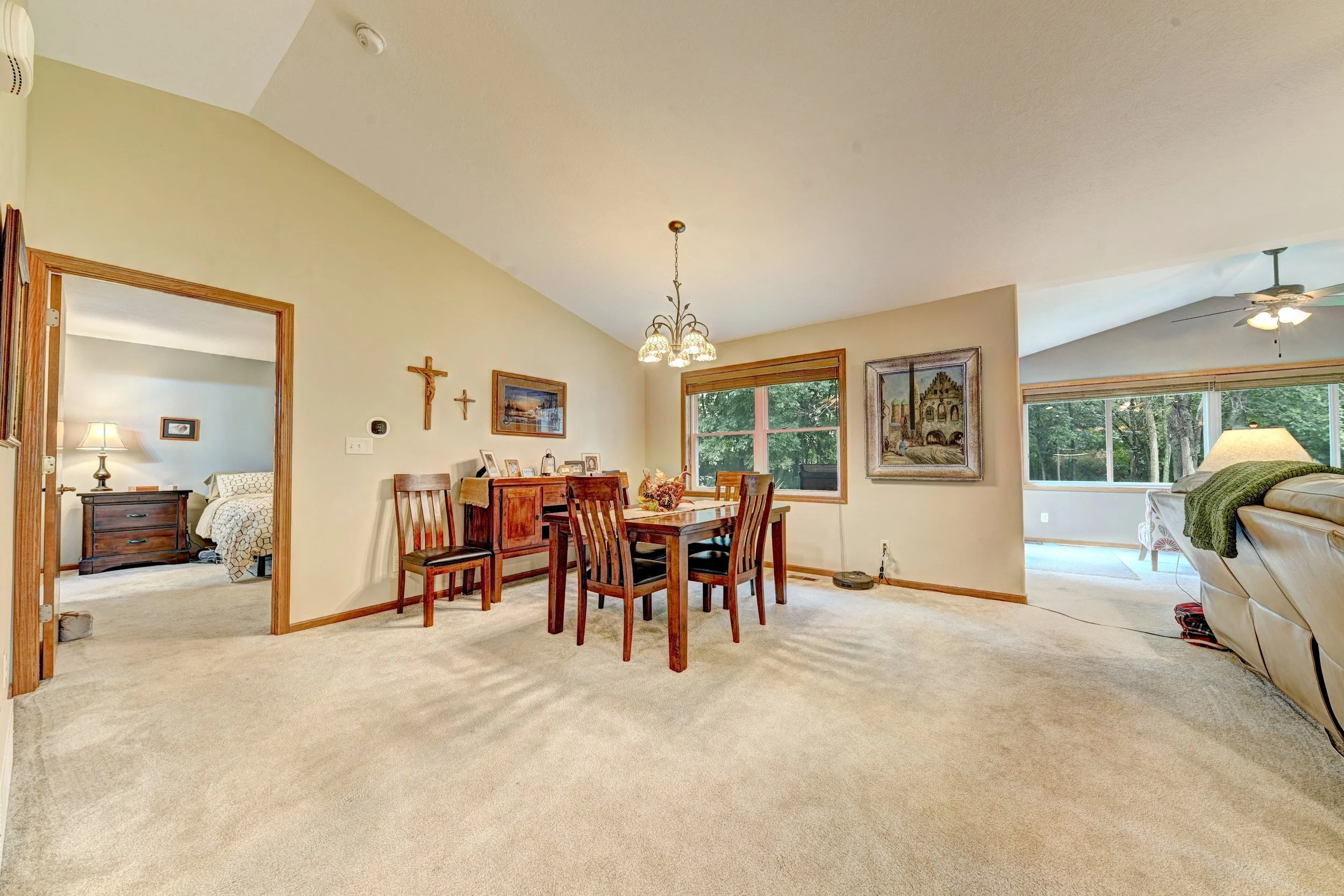 Interior view of a living and dining area with a beige carpet, wooden dining table and chairs, art and cross decorations on the wall, large windows showing green trees outside, and a nearby bedroom with a bed and table lamp.