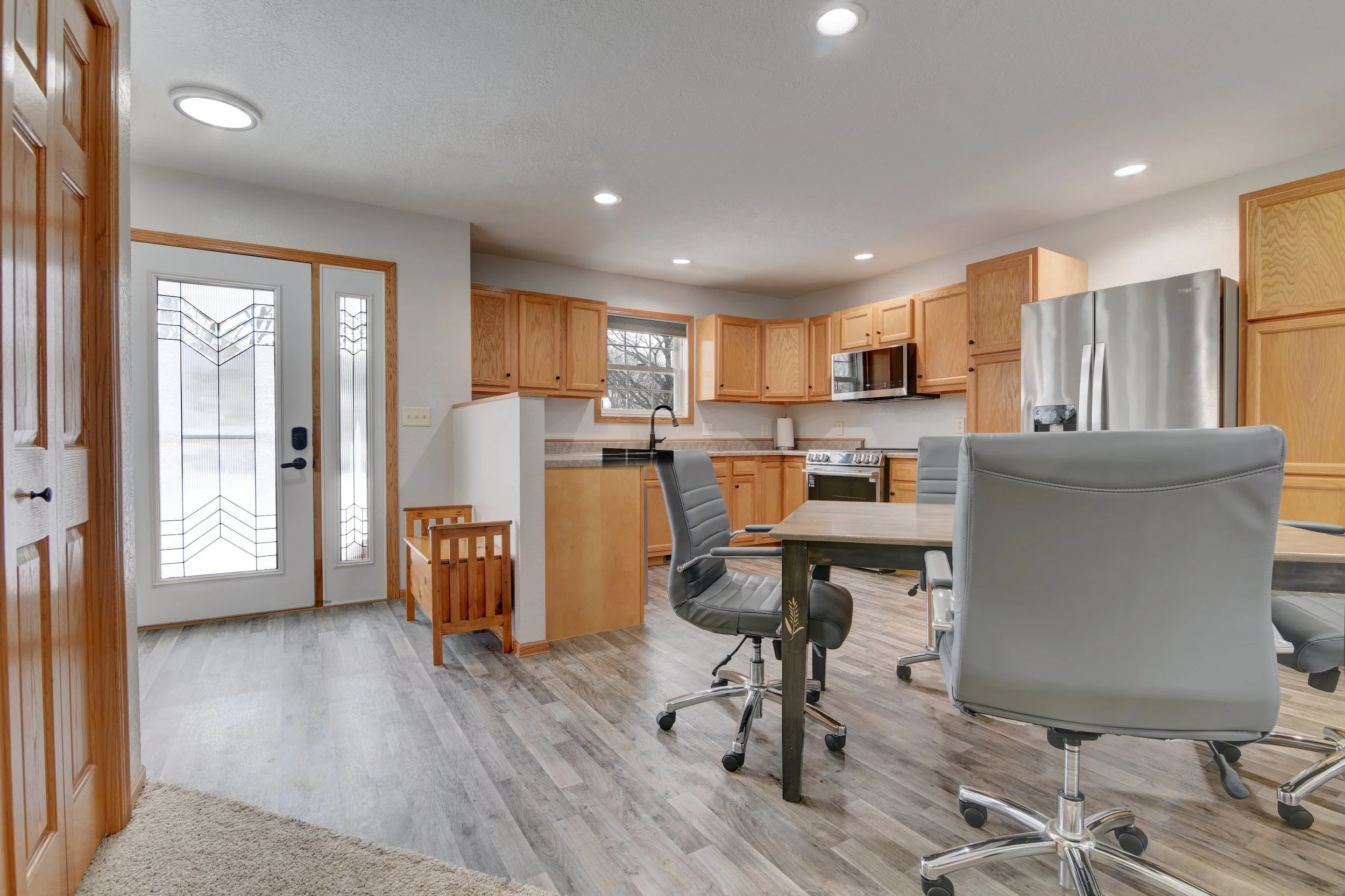 Bright kitchen with wood cabinets, stainless steel refrigerator, microwave, stove, and gray countertop. Open floor plan with a dining area with office chairs around a table, hardwood and carpeted flooring, and a front door with glass panels.
