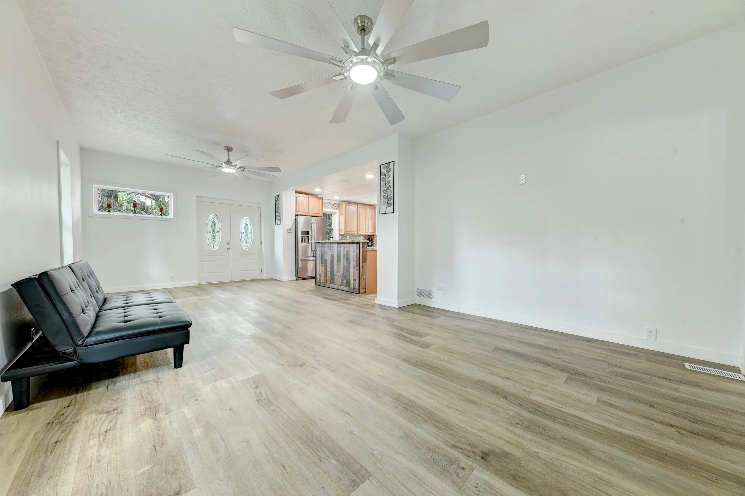 Living room with two ceiling fans, light wood flooring, white walls, a black futon, and a kitchen area with wooden cabinets and stainless steel appliances.