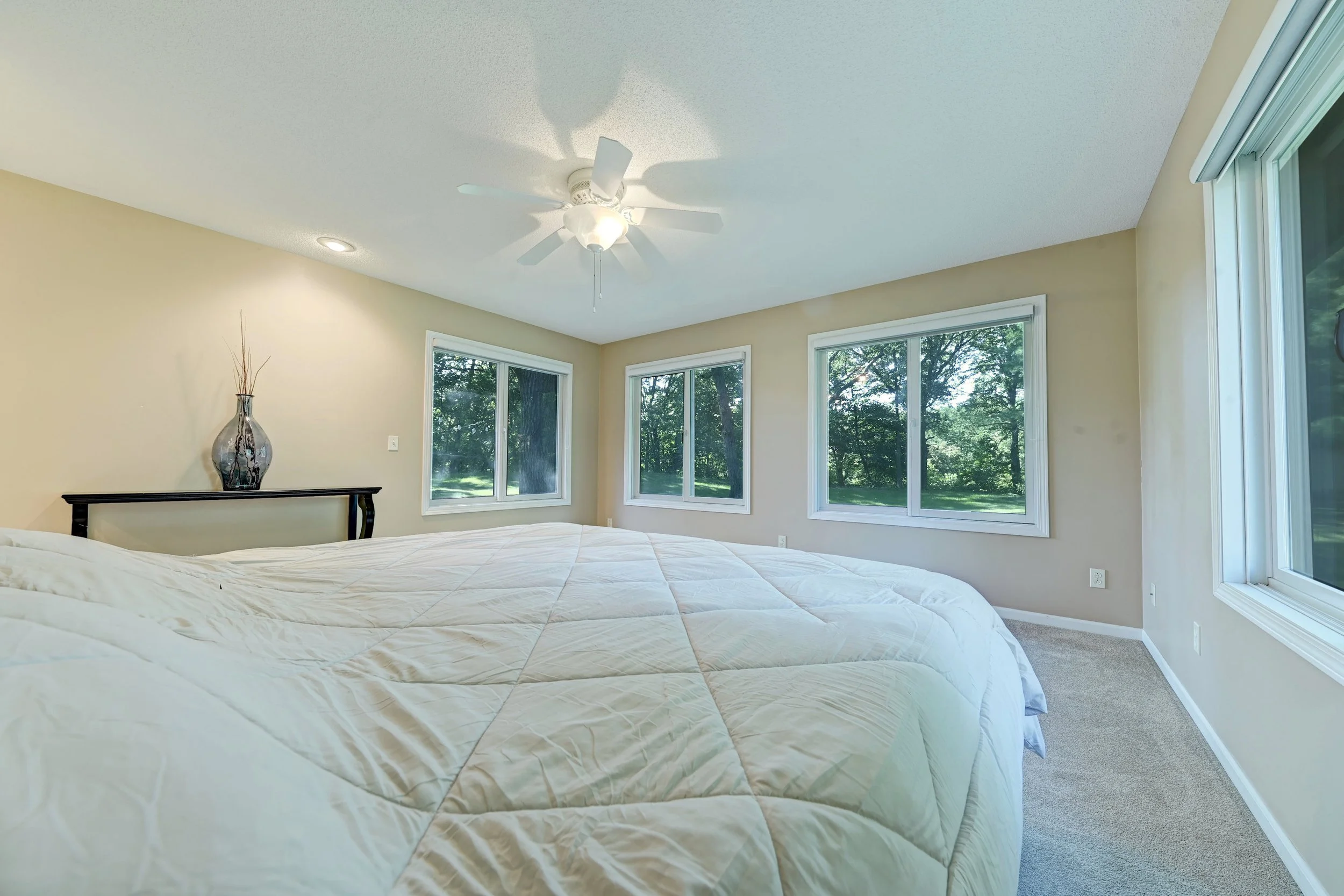 Bright bedroom with a bed, large windows showing greenery outside, a ceiling fan, and a vase with decorative branches on a small table.
